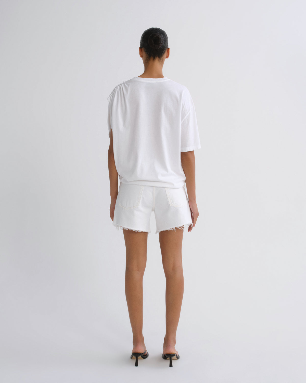 A model viewed from the back wearing a white oversized short-sleeve t-shirt and matching white shorts with frayed hems, paired with black high-heeled sandals, against a light gray studio background.