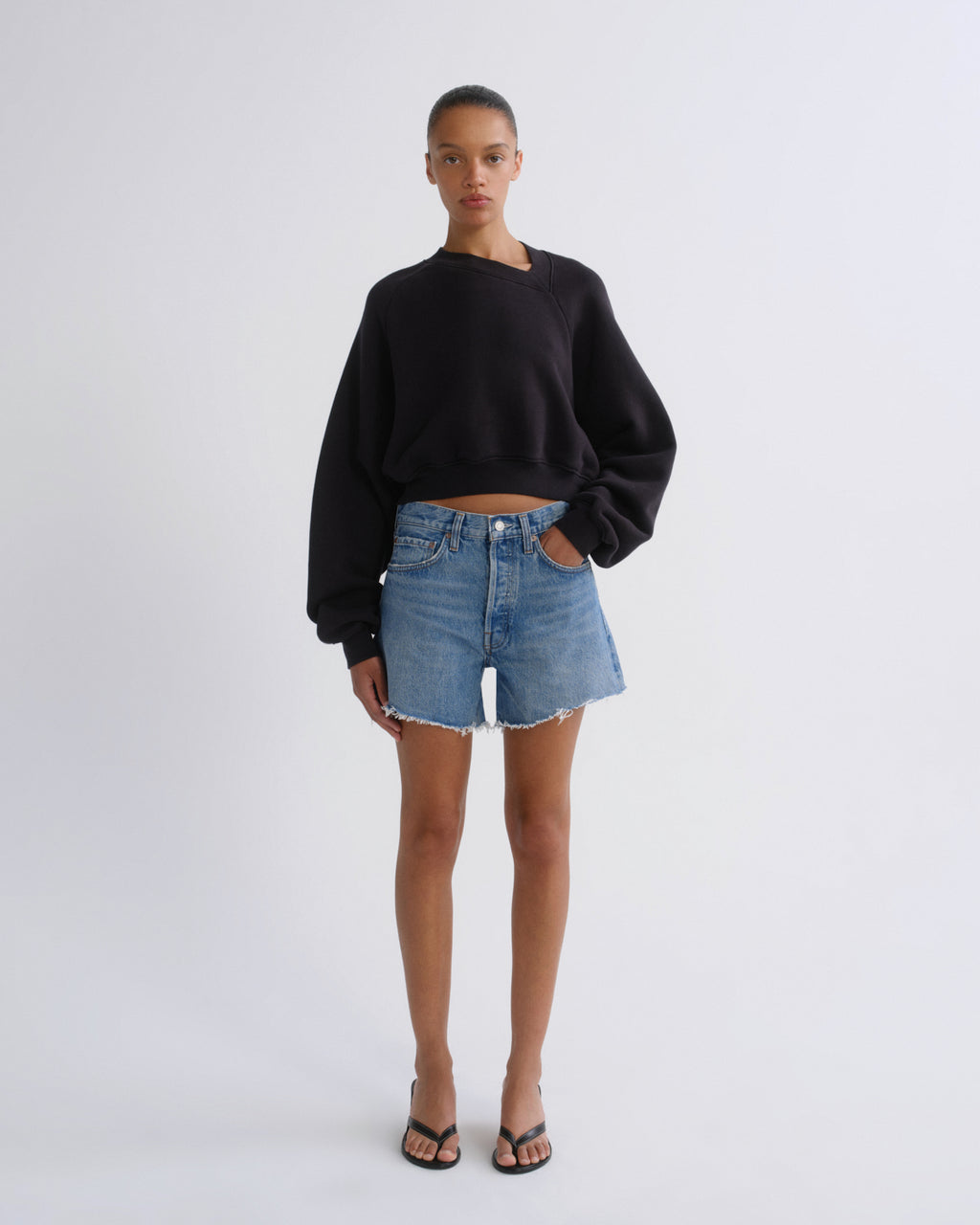 A model stands against a light gray studio background wearing a black asymmetrical long-sleeve crop sweatshirt, light-wash blue denim shorts, and black sandals.
