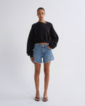 A model stands against a light gray studio background wearing a black asymmetrical long-sleeve crop sweatshirt, light-wash blue denim shorts, and black sandals.