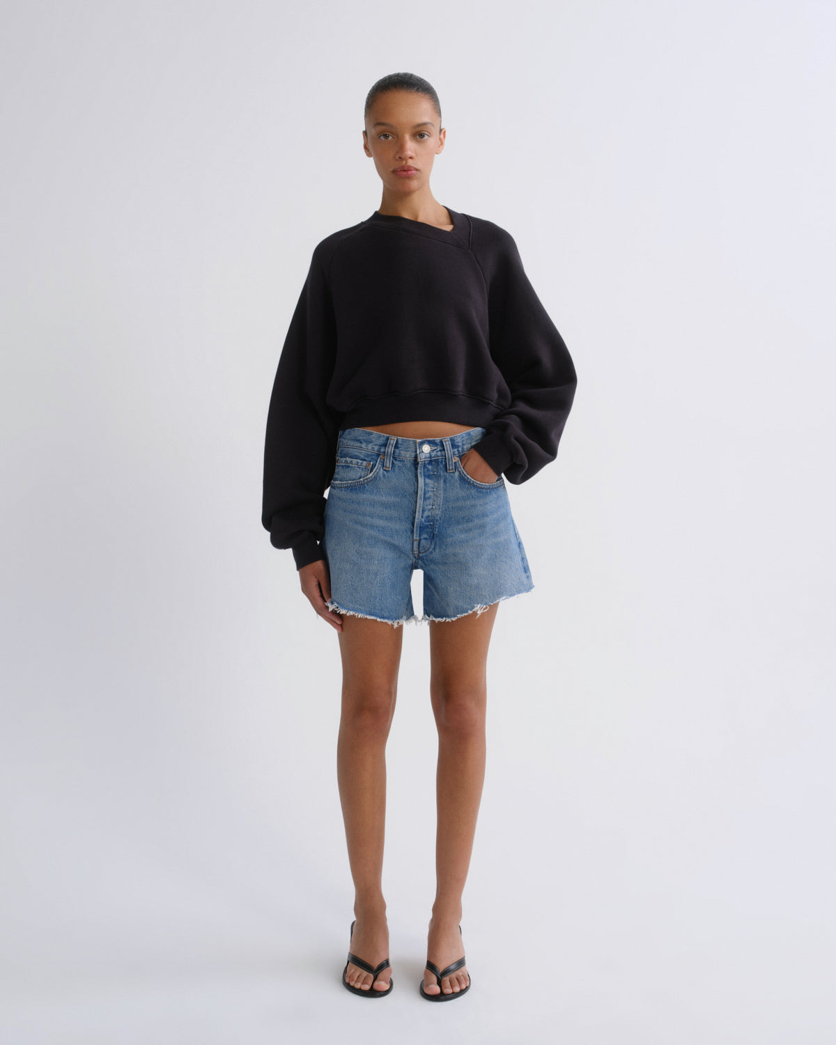 A model stands against a light gray studio background wearing a black asymmetrical long-sleeve crop sweatshirt, light-wash blue denim shorts, and black sandals.