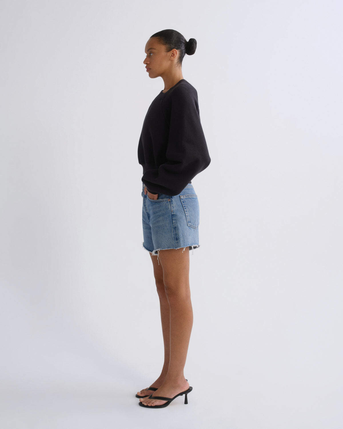 A woman stands in profile wearing a black crewneck sweatshirt, light-wash denim shorts, and black heeled sandals against a plain light gray studio background.