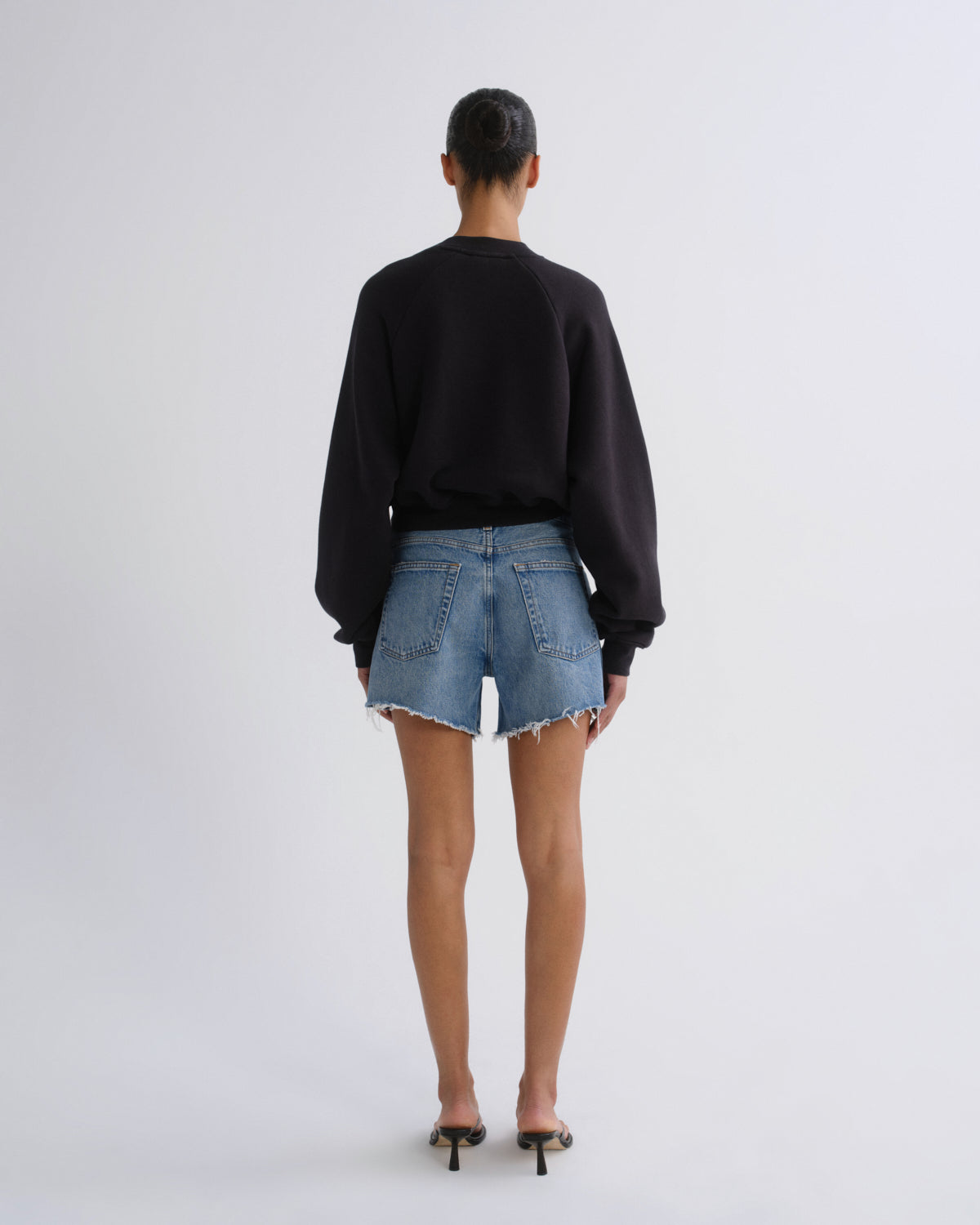 A model viewed from behind wearing a black long-sleeve crewneck sweatshirt, light-wash blue denim shorts with a raw hem, and black high-heeled sandals, set against a plain light gray studio background.