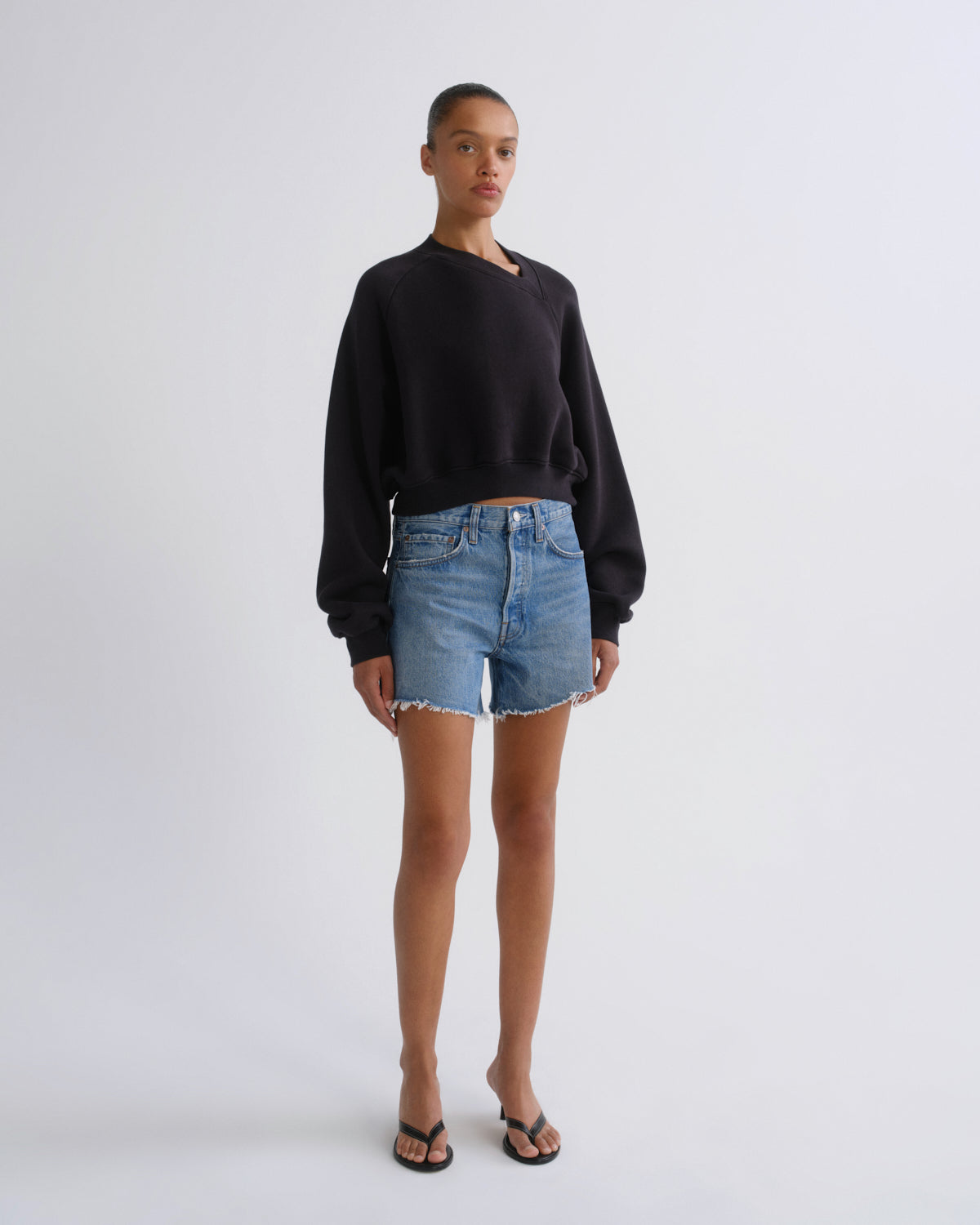 A model stands against a light gray studio background wearing a black long-sleeve crewneck sweatshirt, light-wash denim shorts with a raw hem, and black flip-flop sandals.