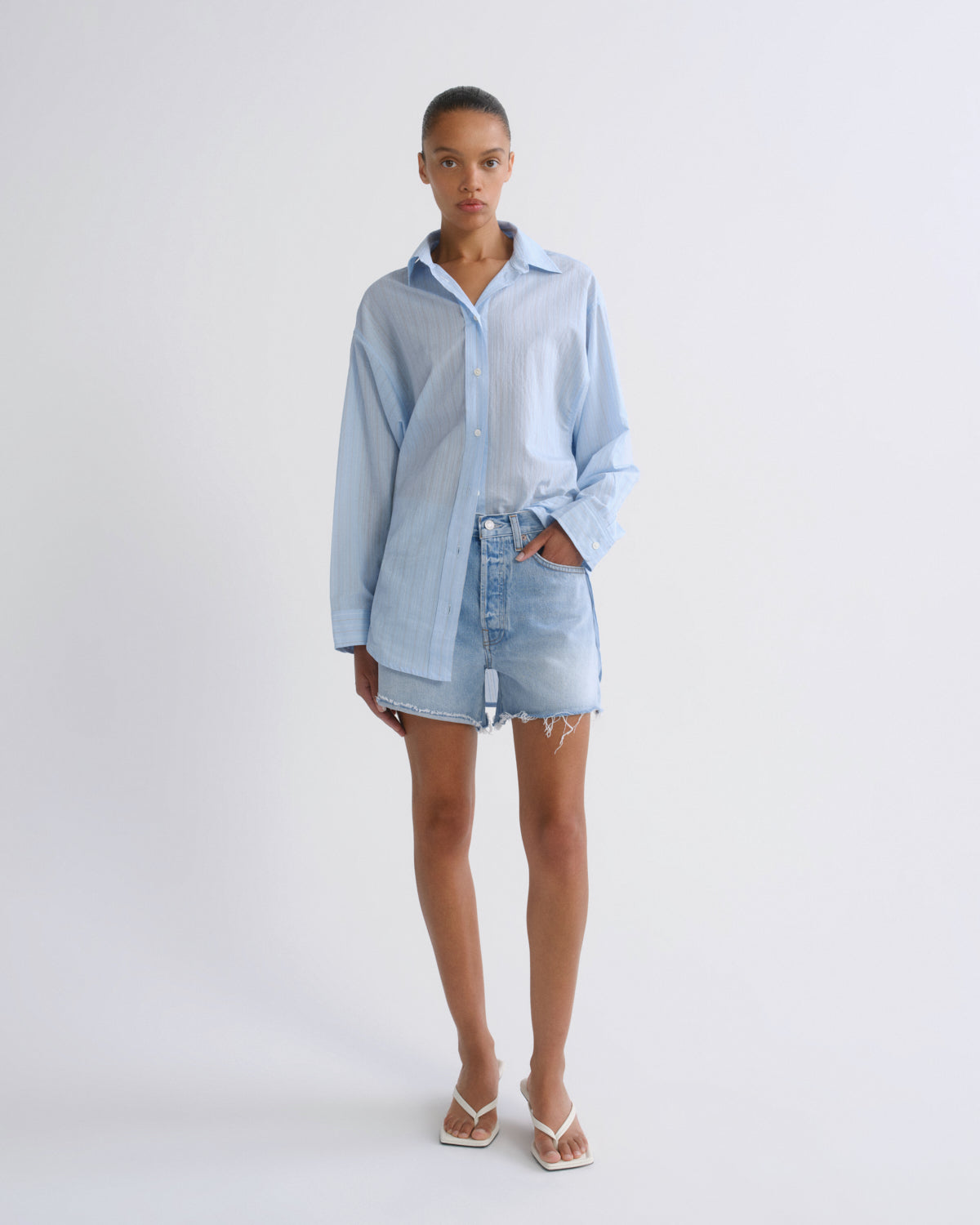 A model stands against a plain light gray studio background wearing a light blue oversized button-up shirt, matching light blue denim shorts, and white thong sandals.