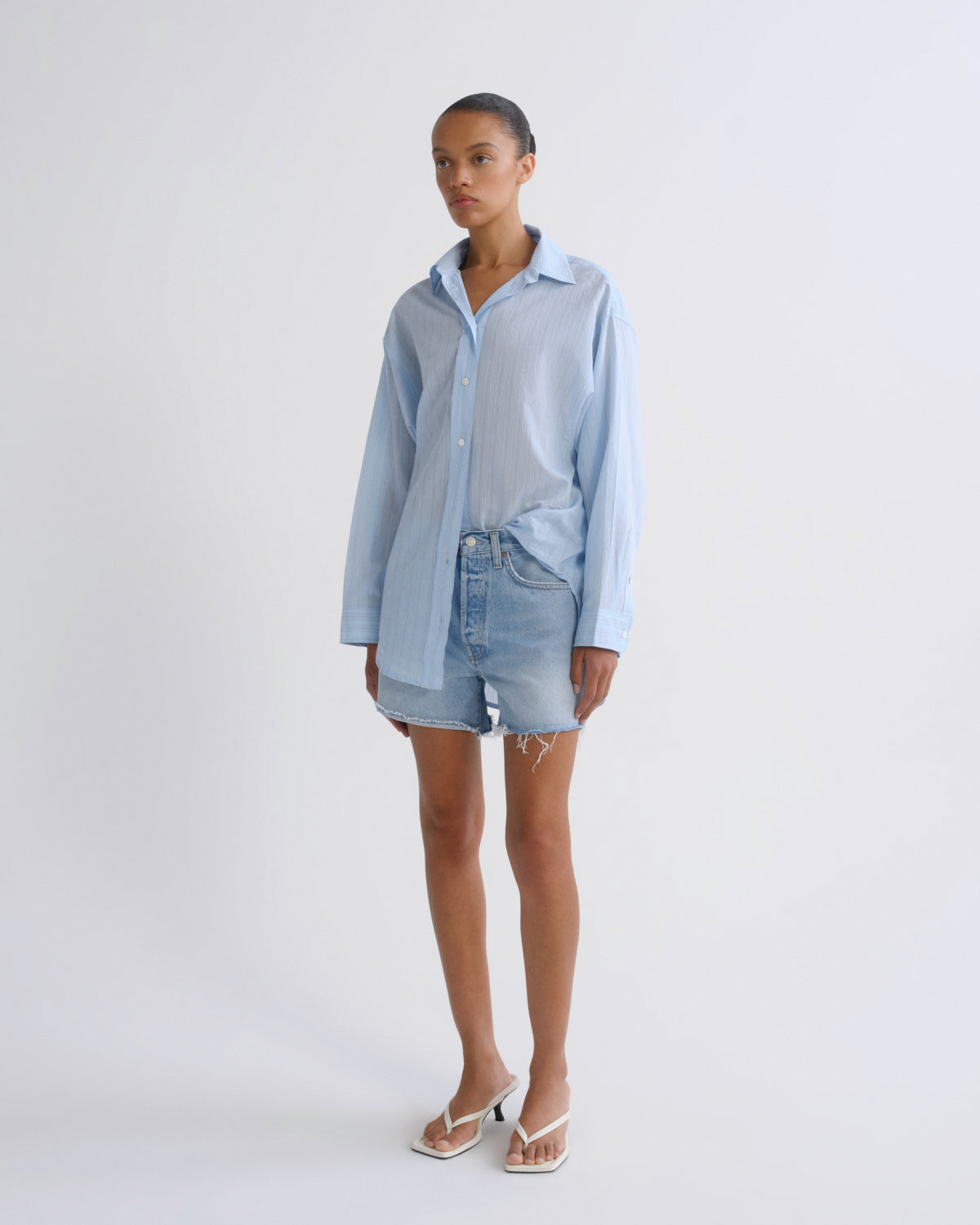 A model stands against a light gray studio background wearing a light blue striped button-up shirt, light-wash denim shorts, and white heeled sandals.