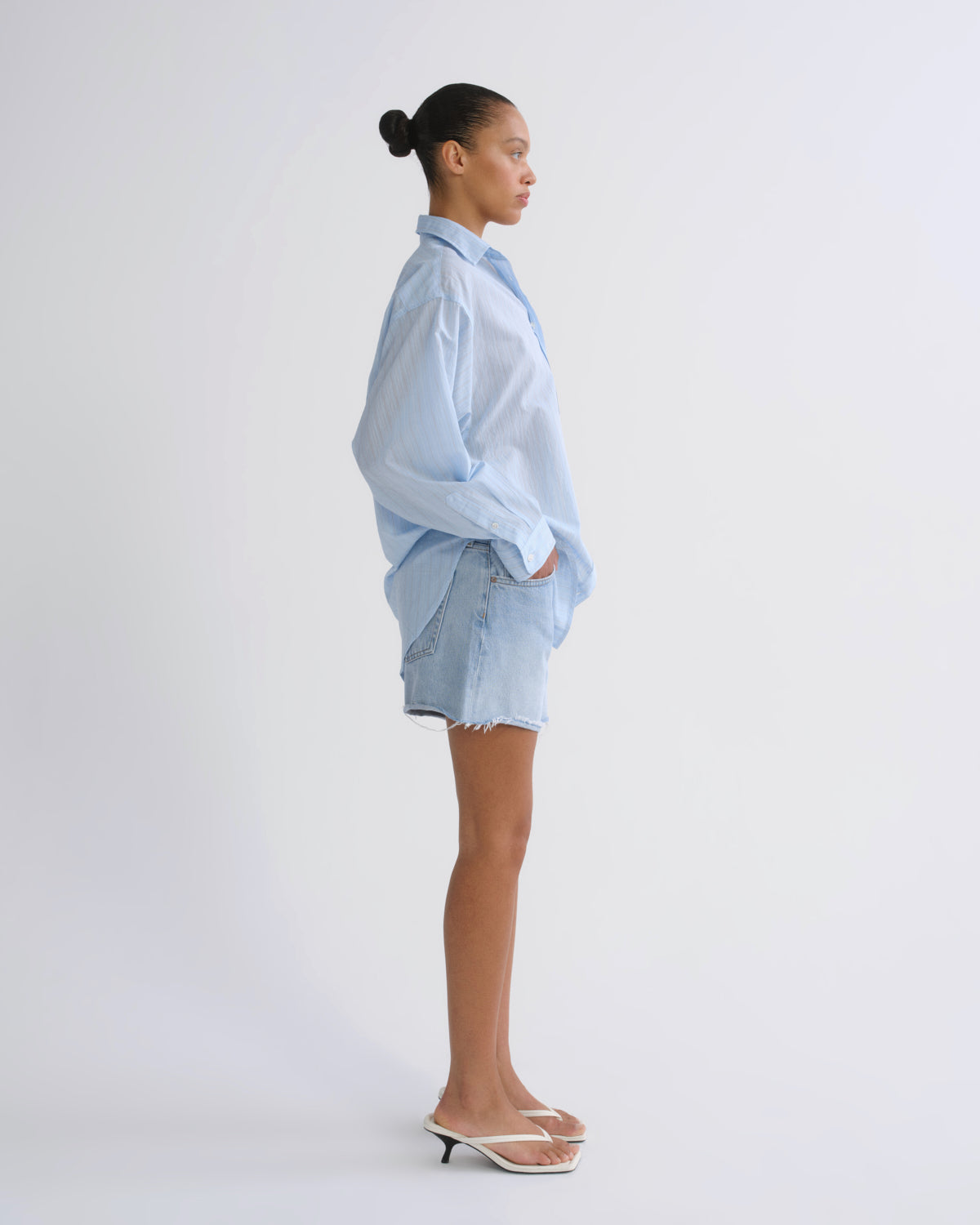 A model shown in profile wearing a light blue oversized button-down shirt, light-wash denim shorts, and white slide sandals against a plain light gray studio background.