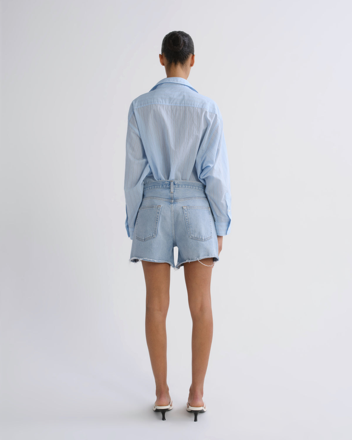 A model viewed from behind wearing a light blue denim shirt and matching denim shorts, paired with white heeled sandals, against a plain light gray studio background.