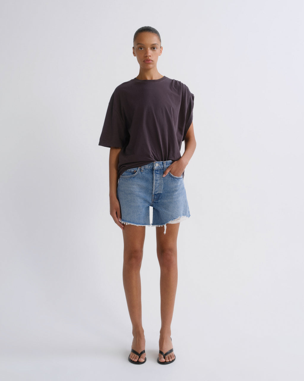 A model stands against a light gray studio background wearing an oversized dark charcoal t-shirt, light-wash denim shorts, and black flip-flops.