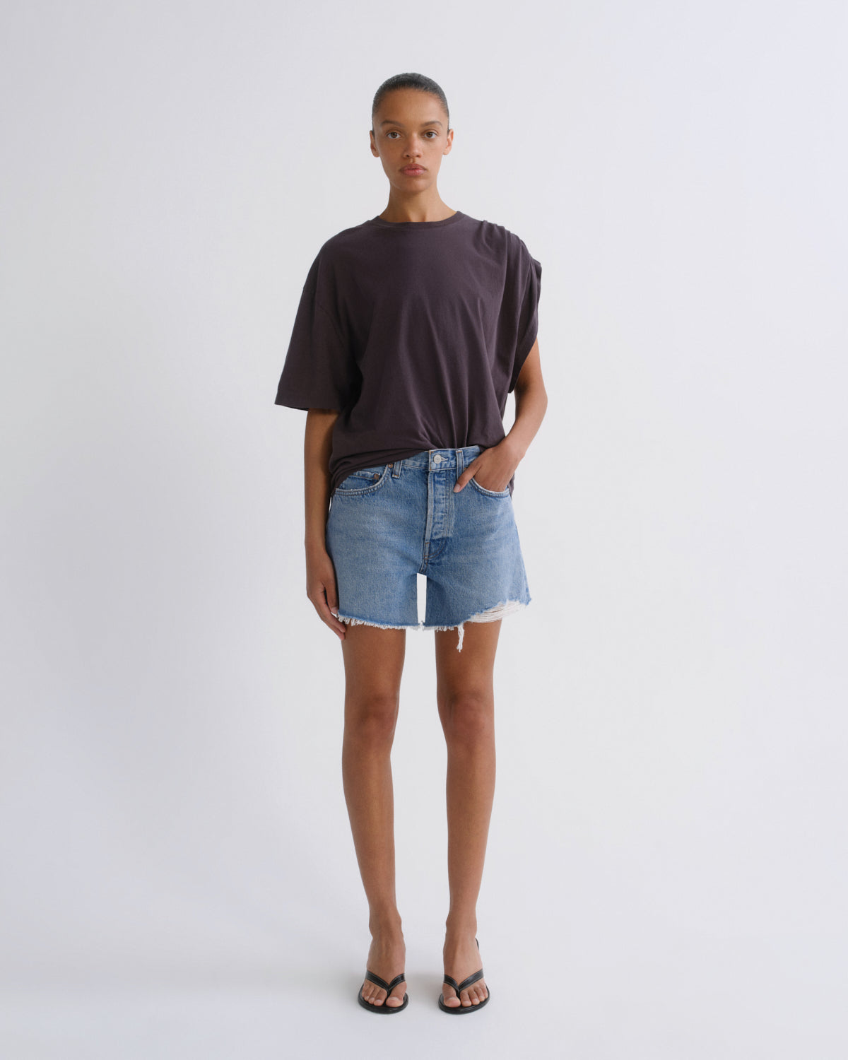 A model stands against a light gray studio background wearing an oversized dark charcoal t-shirt, light-wash denim shorts, and black flip-flops.