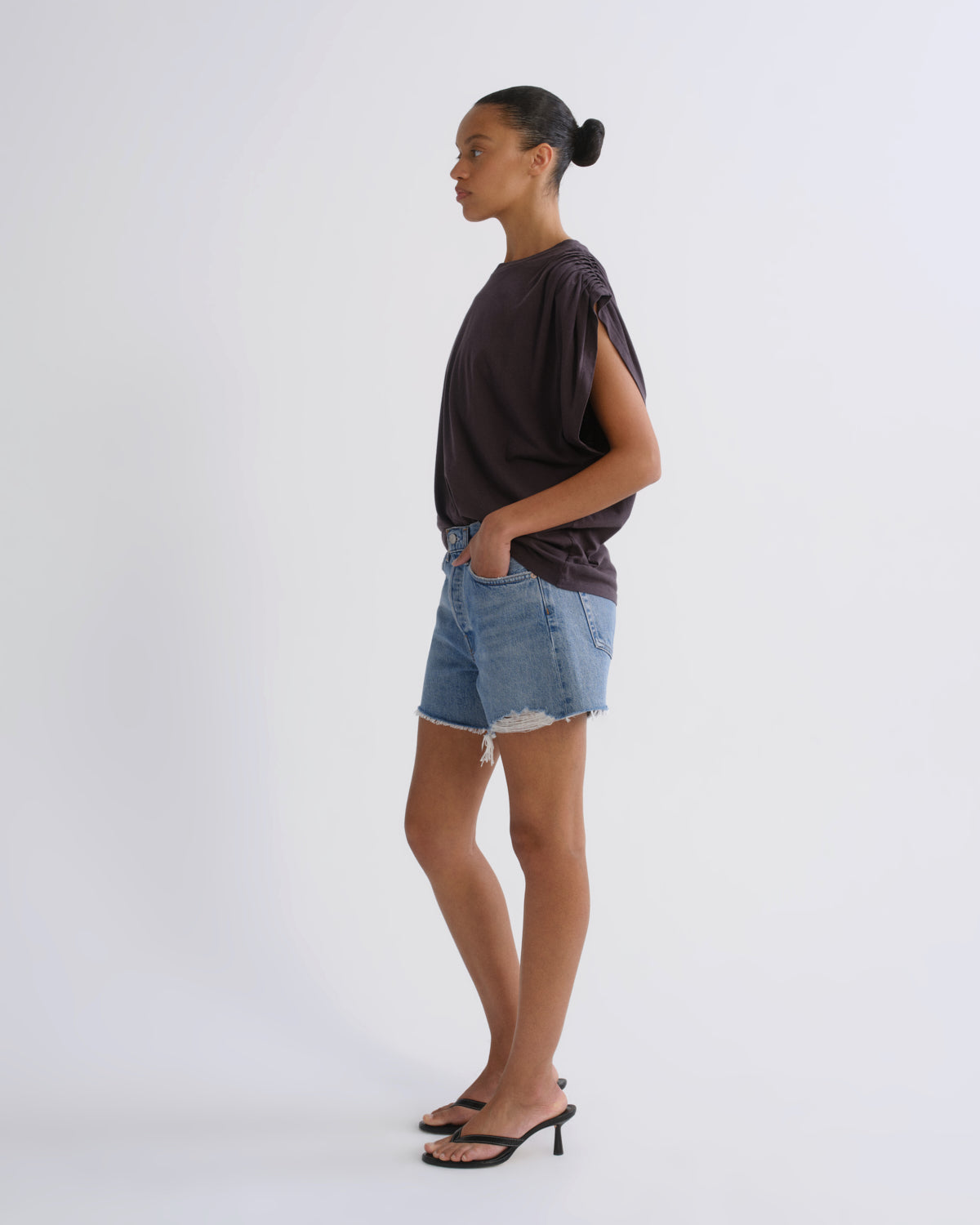 A woman stands in profile wearing a dark brown short-sleeve top, light-wash denim shorts, and black heeled sandals against a plain light gray studio background.