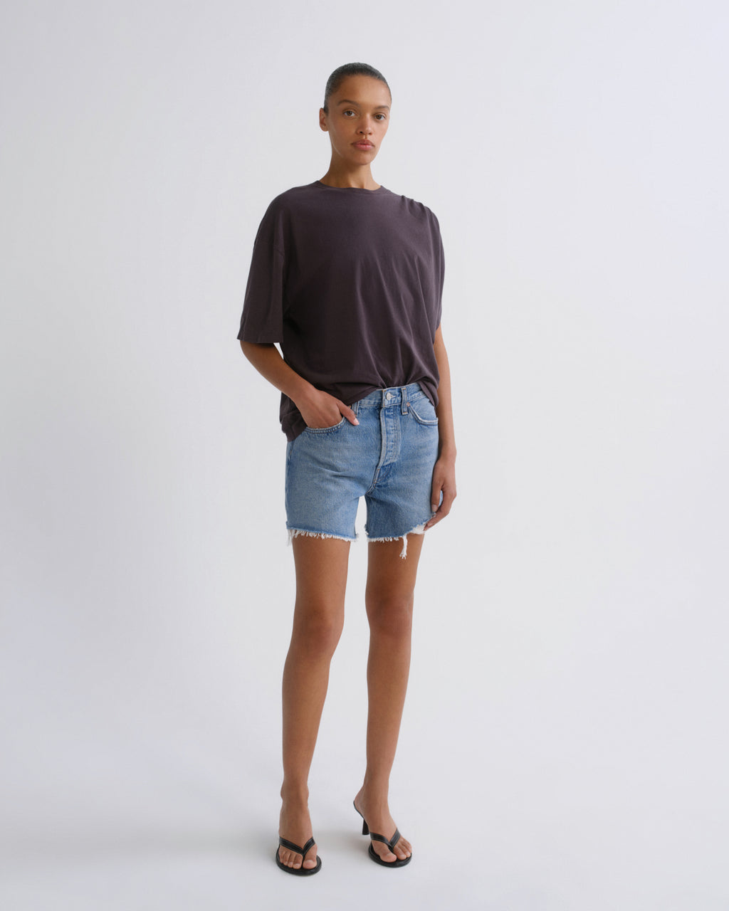 A model stands against a light gray studio background wearing a dark charcoal t-shirt, light-wash denim shorts with a raw hem, and black flip-flop sandals.