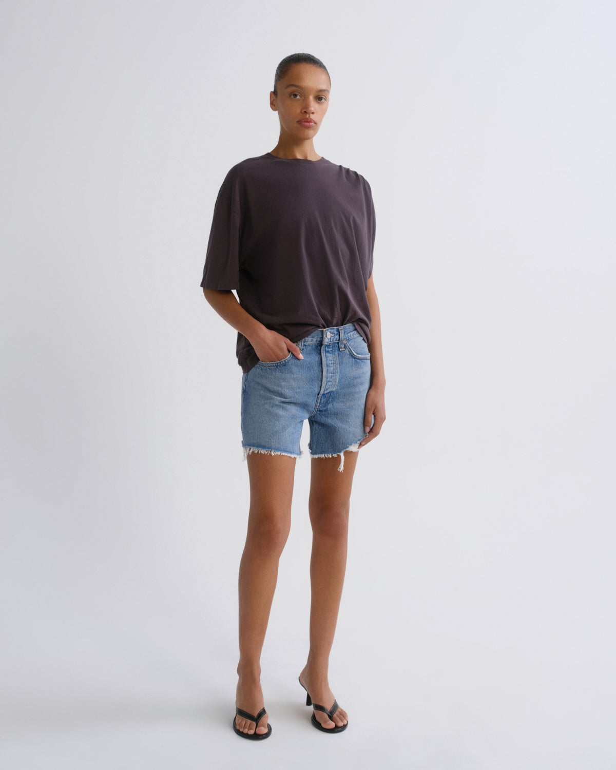 A model stands against a light gray studio background wearing a dark charcoal t-shirt, light-wash denim shorts with a raw hem, and black flip-flop sandals.