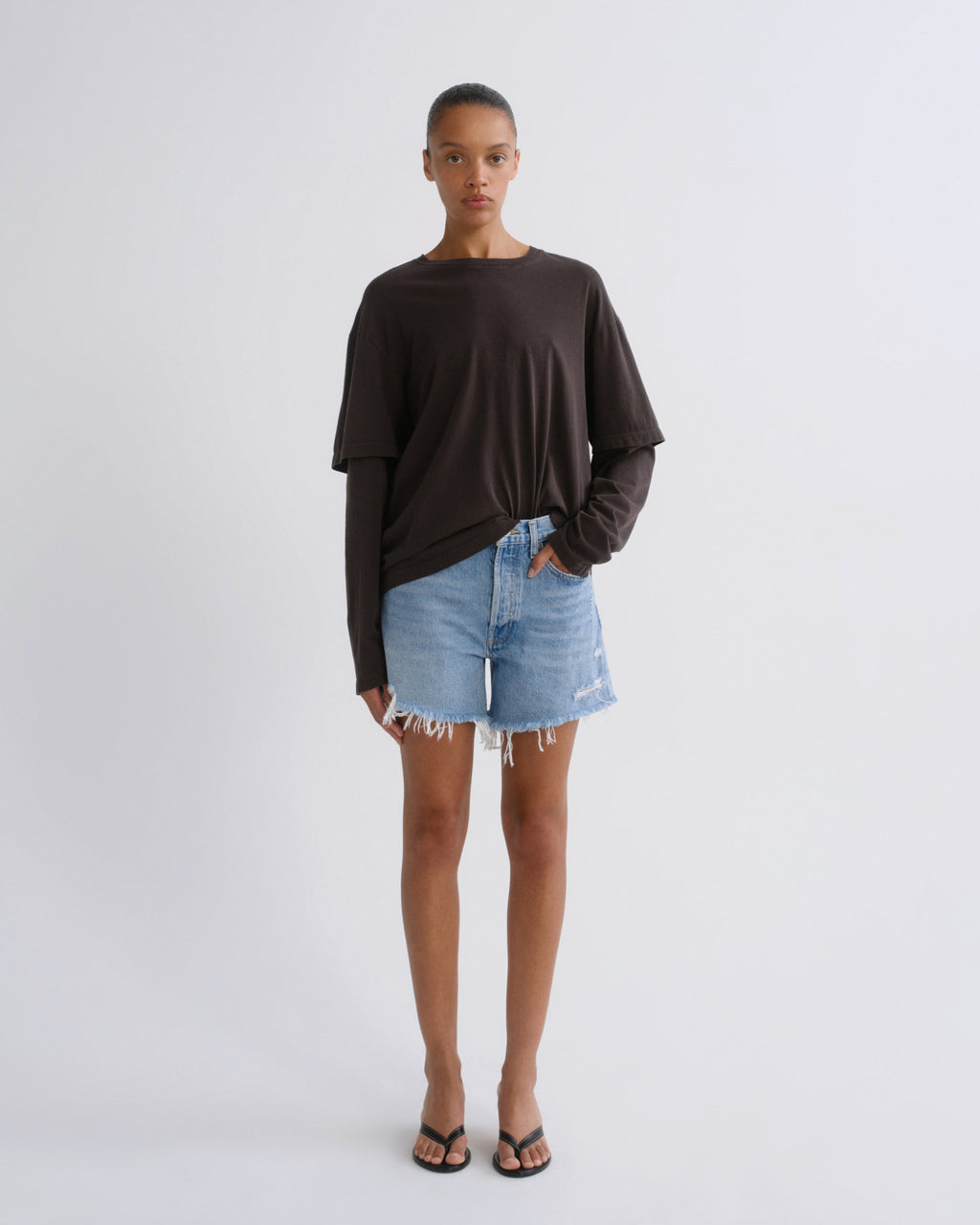 A model stands against a light gray studio background wearing a dark brown long-sleeve top, light blue denim shorts, and black flip-flop sandals.