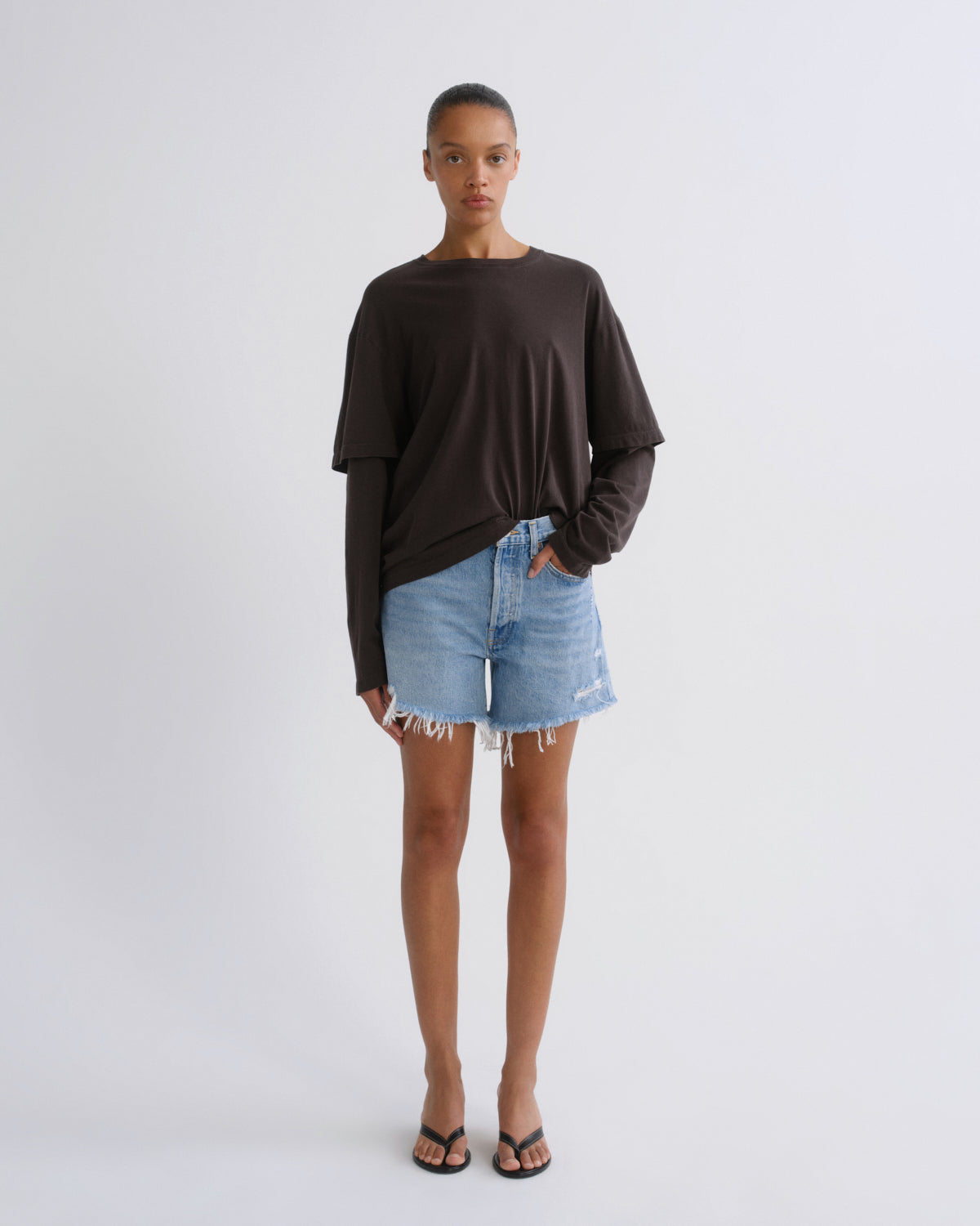 A model stands against a light gray studio background wearing a dark brown long-sleeve top, light blue denim shorts, and black flip-flop sandals.