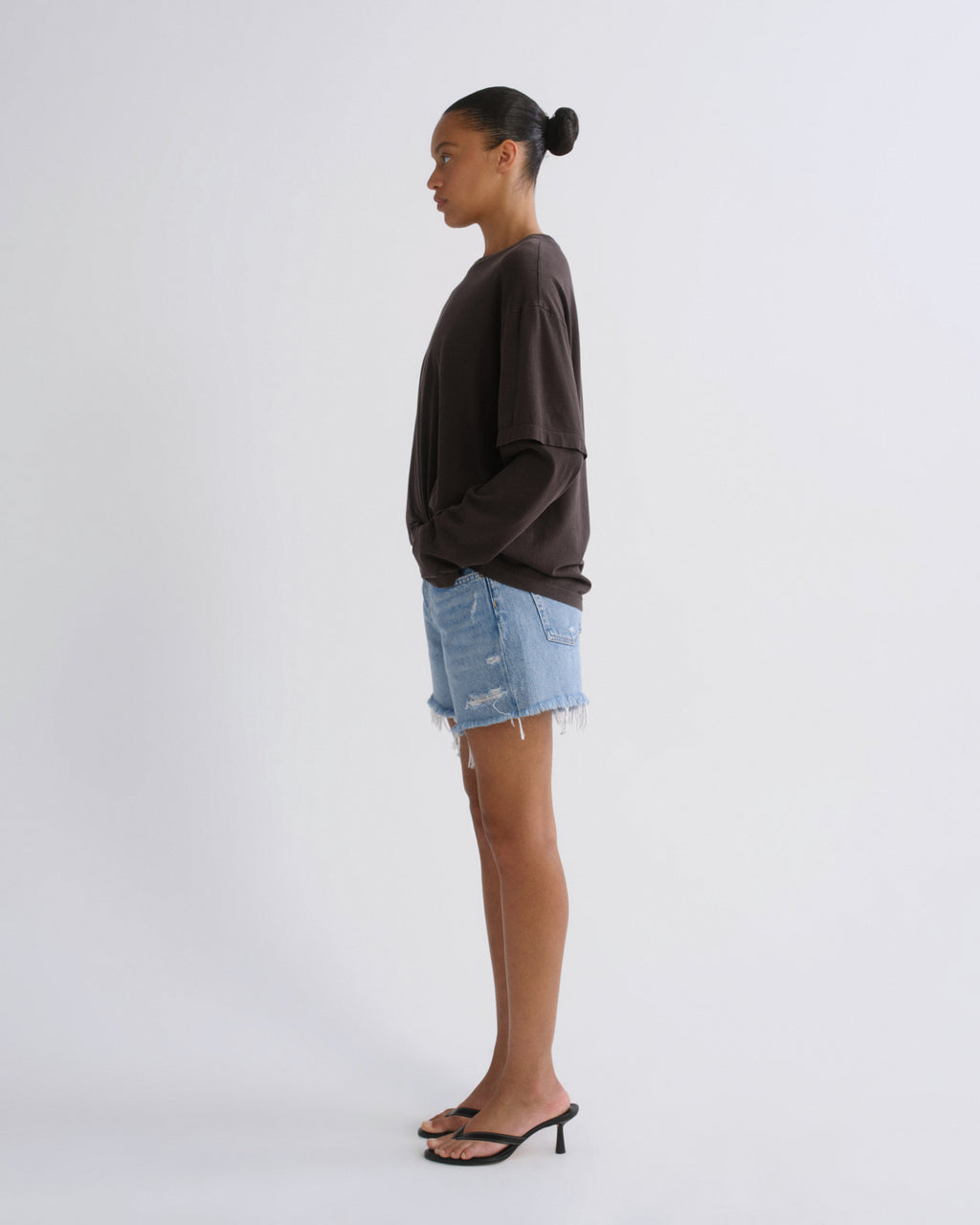 A woman stands in profile wearing a dark brown oversized long-sleeve top, light blue distressed denim shorts, and black heeled sandals against a plain light gray studio background.