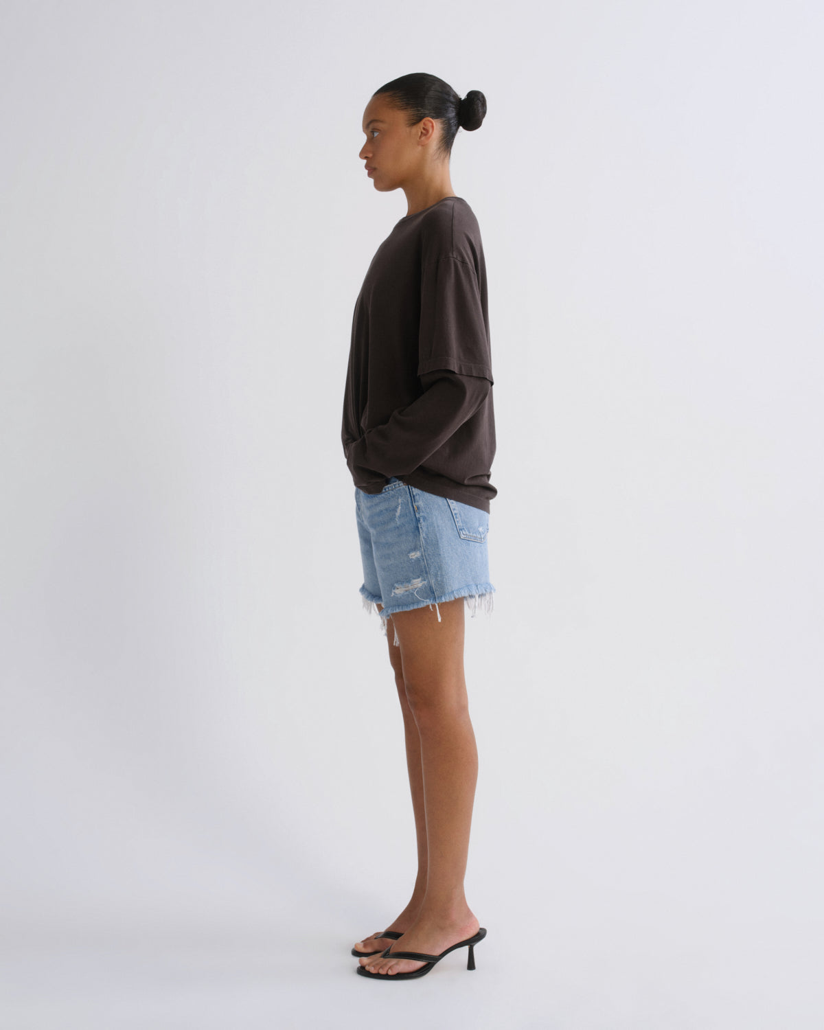 A woman stands in profile wearing a dark brown oversized long-sleeve top, light blue distressed denim shorts, and black heeled sandals against a plain light gray studio background.