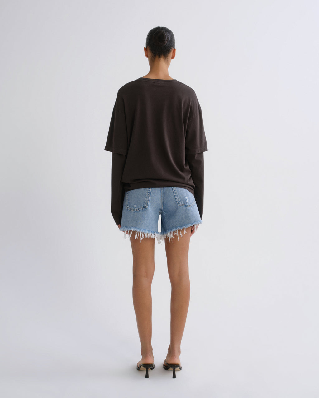 A model viewed from behind wearing an oversized dark brown long-sleeve top, light-wash denim shorts with frayed hems, and black heeled sandals against a plain light gray studio background.