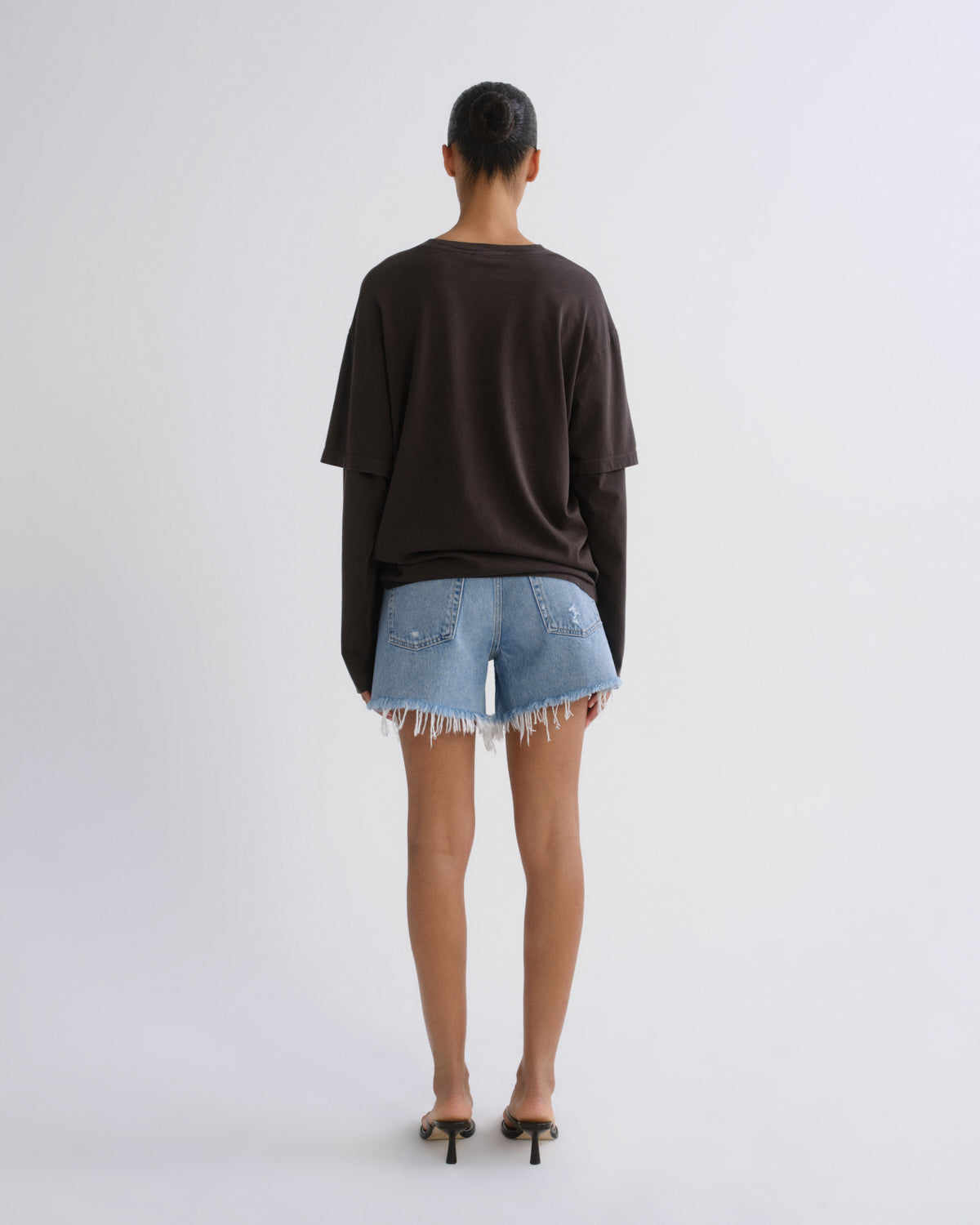 A model viewed from behind wearing an oversized dark brown long-sleeve top, light-wash denim shorts with frayed hems, and black heeled sandals against a plain light gray studio background.
