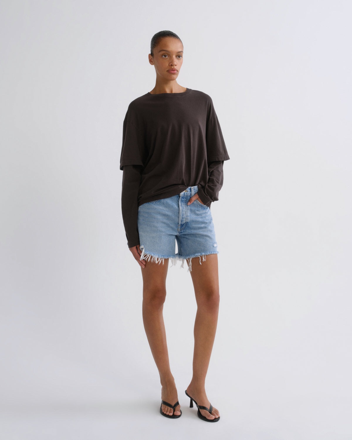 A model stands against a light gray studio background wearing a dark brown long-sleeve t-shirt, light-wash denim shorts with frayed hems, and black thong sandals.