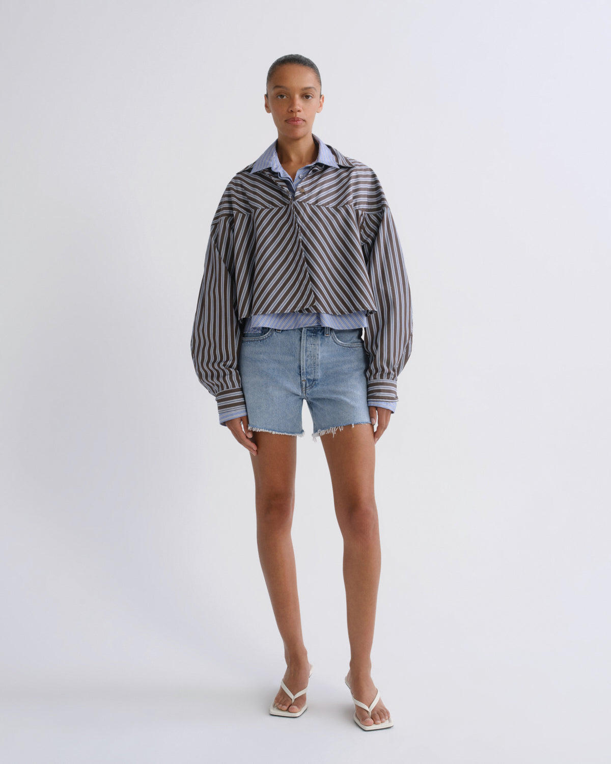 A model stands against a light gray studio background wearing a cropped long-sleeve button-up shirt with vertical black and white stripes, light-wash denim shorts, and white thong sandals.