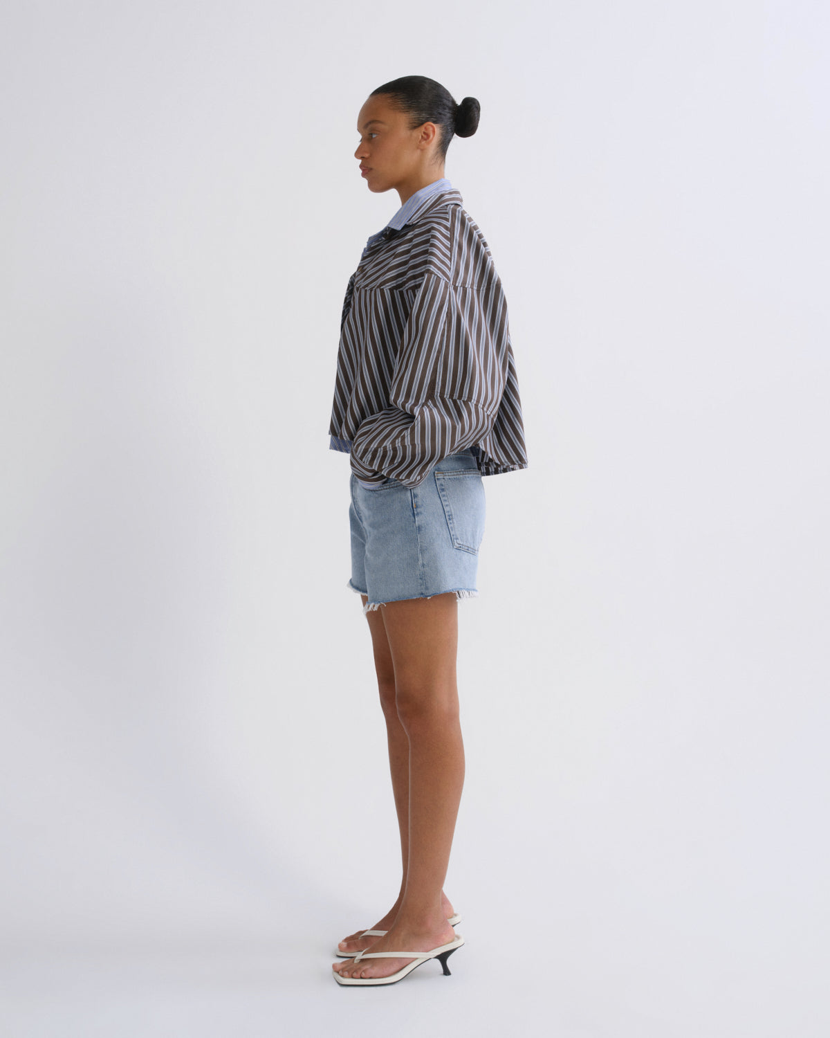 A model is shown in profile wearing a black and white vertically striped button-up shirt, light-wash denim shorts, and white heeled sandals against a plain white studio background.