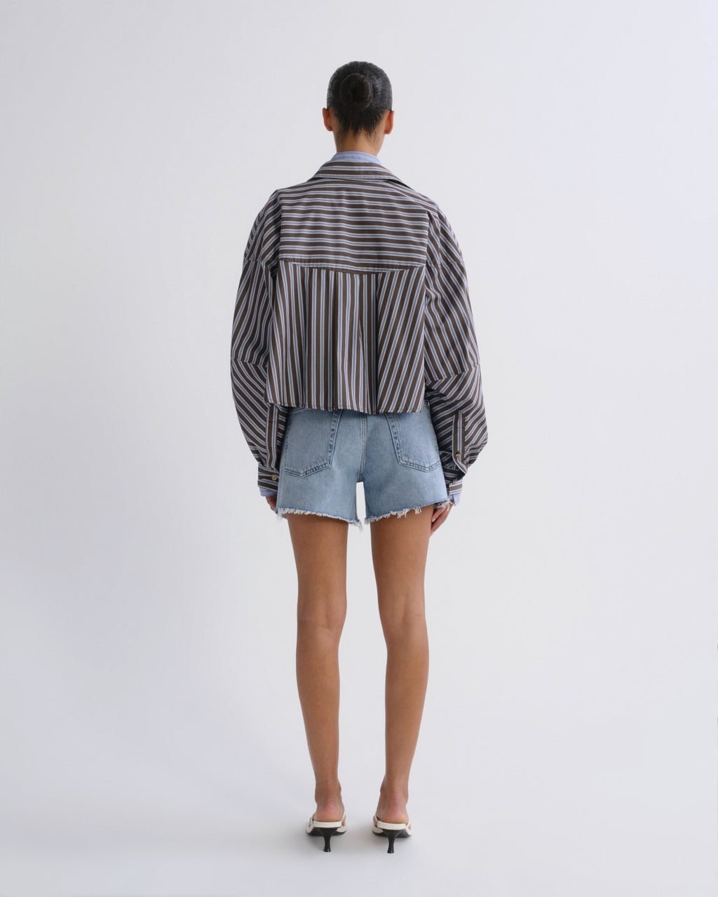A model viewed from the back wearing a cropped, long-sleeve striped button-up shirt in black and white, paired with light-wash denim shorts and white heeled sandals, against a plain white studio background.