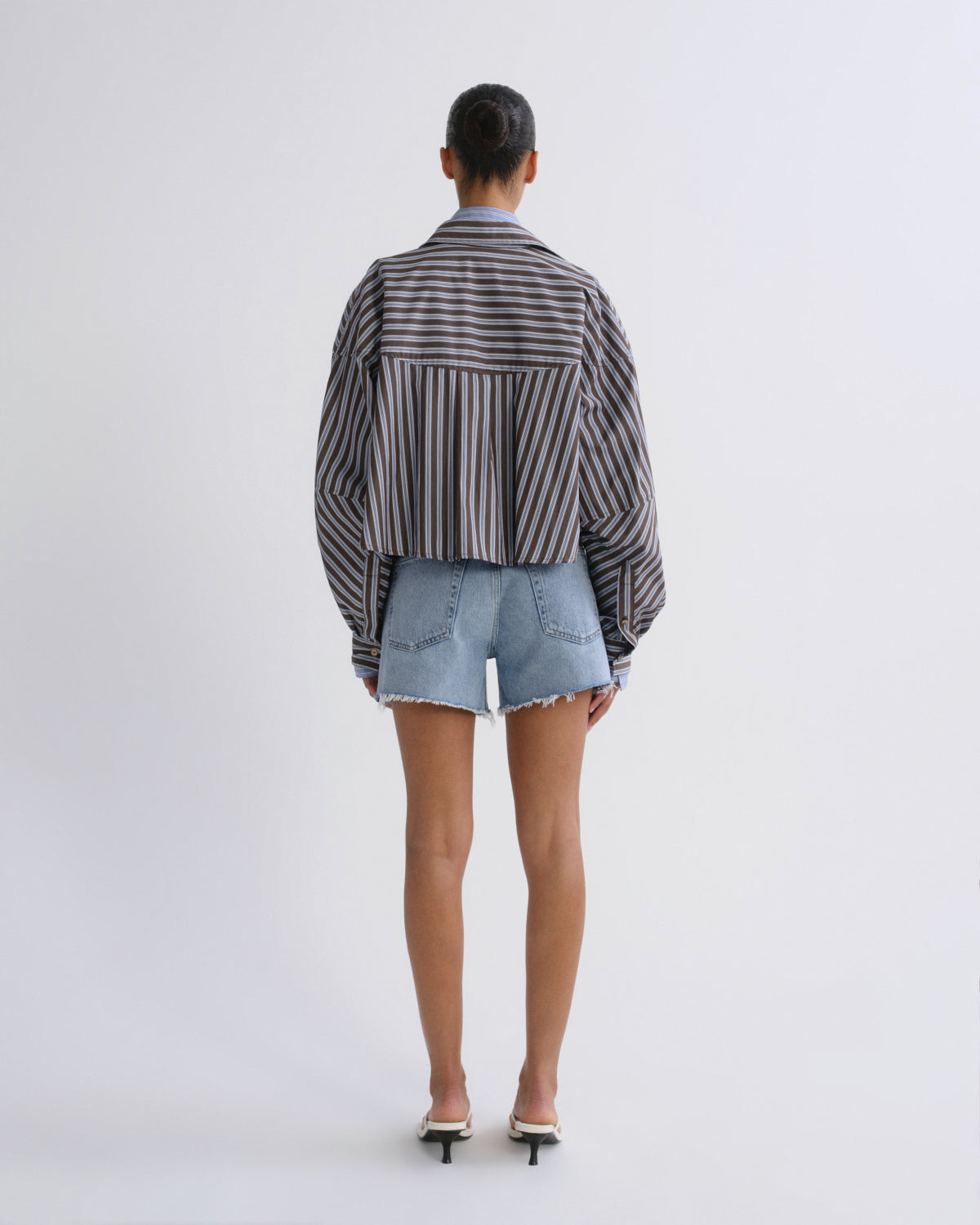 A model viewed from the back wearing a cropped, long-sleeve striped button-up shirt in black and white, paired with light-wash denim shorts and white heeled sandals, against a plain white studio background.