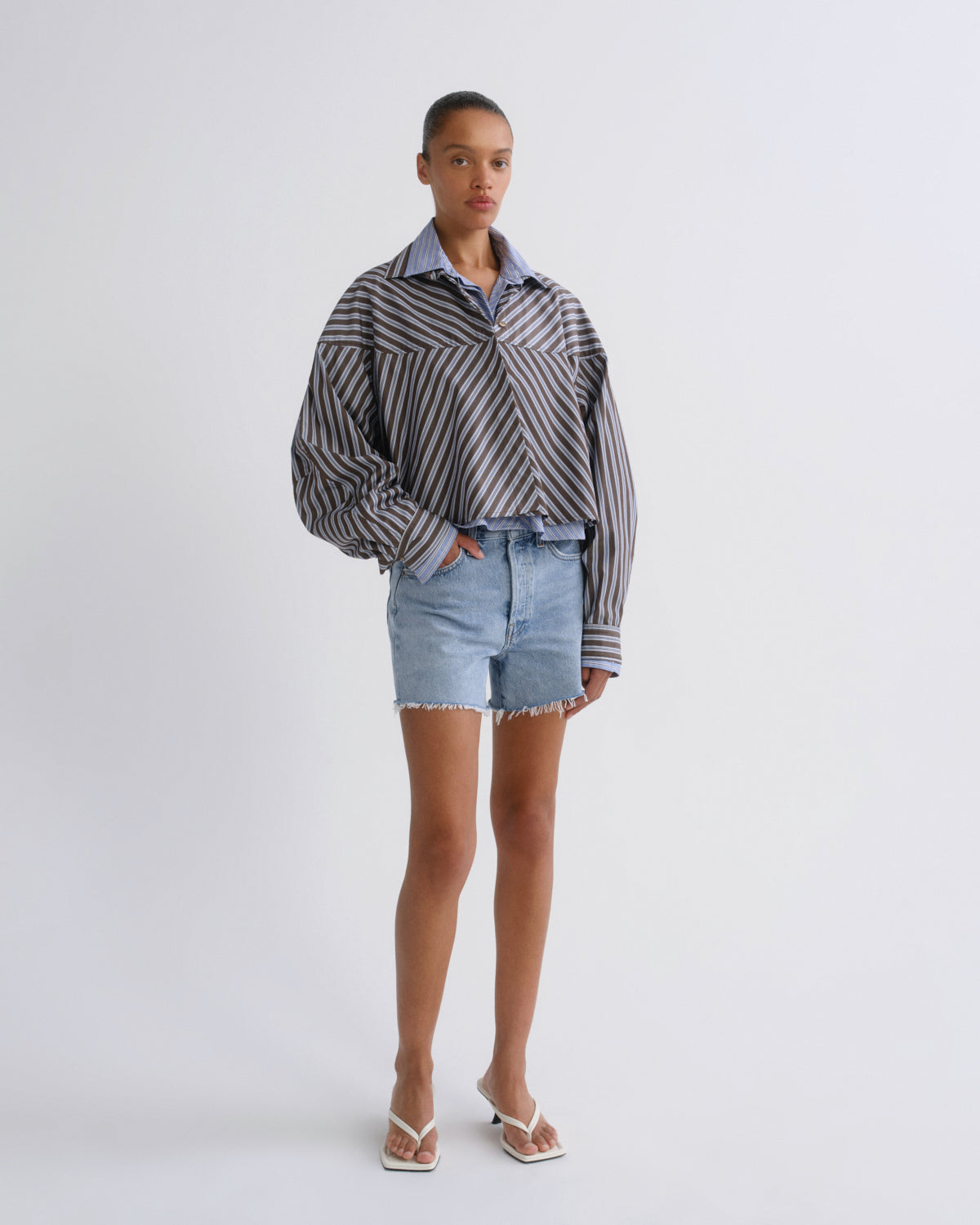 A model stands against a light gray studio background wearing a striped button-up shirt with a light blue collared shirt underneath, light-wash denim shorts, and white flip-flop sandals.
