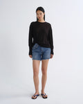 A woman stands against a light gray studio background wearing a loose black long-sleeve top, blue denim shorts, and blue flip-flop sandals.