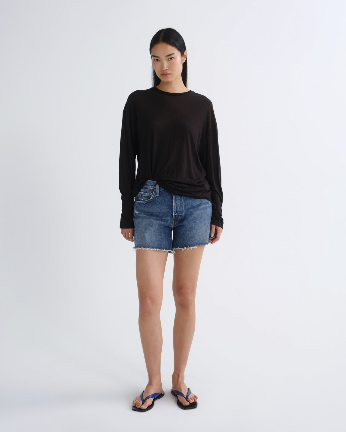 A woman stands against a light gray studio background wearing a loose black long-sleeve top, blue denim shorts, and blue flip-flop sandals.