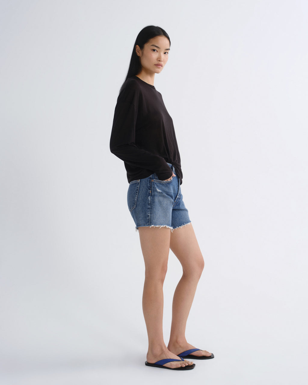 A woman stands in profile wearing a black long-sleeve top, blue denim shorts, and dark blue flip-flops against a plain light gray studio background.