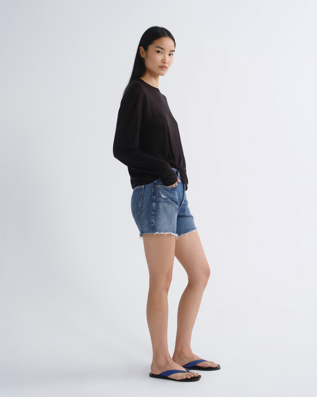 A woman stands in profile wearing a black long-sleeve top, blue denim shorts, and dark blue flip-flops against a plain light gray studio background.
