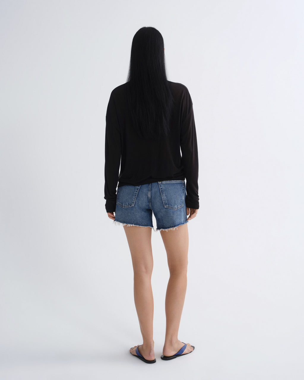 A woman with long black hair is shown from behind wearing a black long-sleeve top, blue denim shorts, and black slide sandals against a plain light gray studio background.