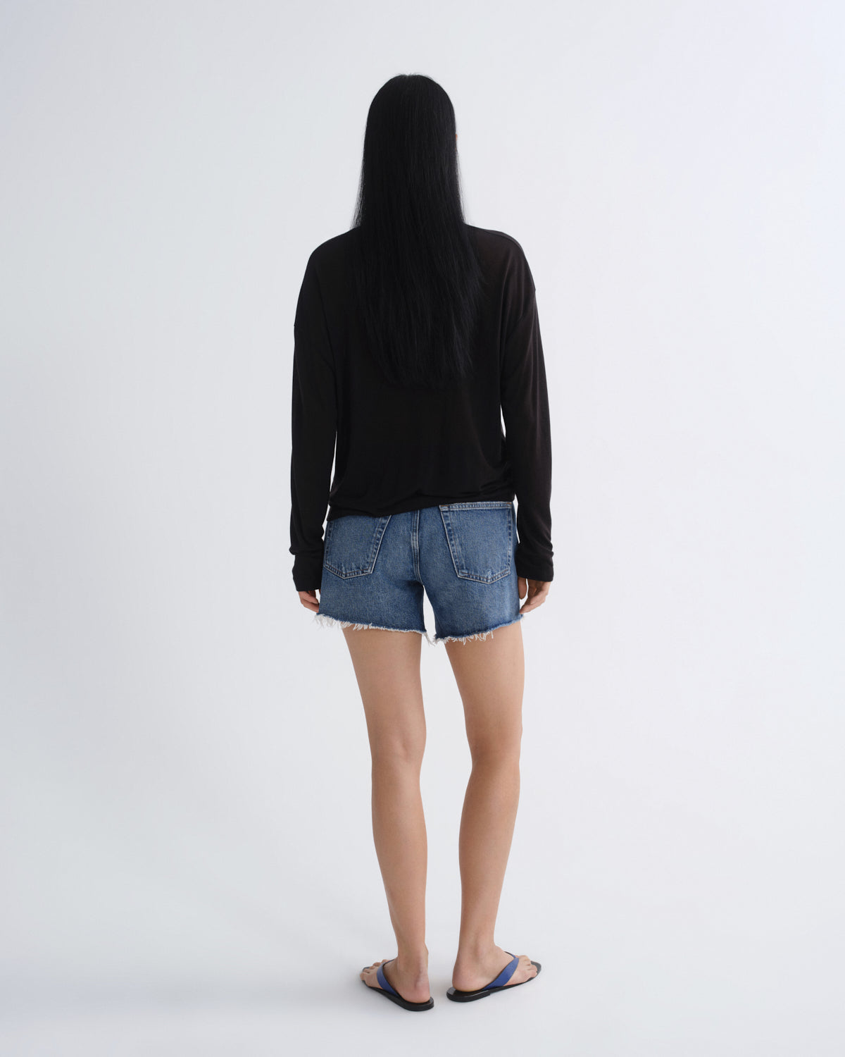 A woman with long black hair is shown from behind wearing a black long-sleeve top, blue denim shorts, and black slide sandals against a plain light gray studio background.