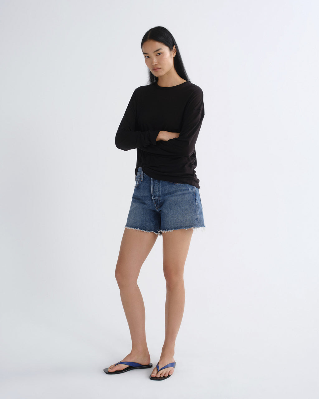 A woman modeling a black long-sleeve crewneck top, blue denim shorts, and blue flip-flop sandals against a plain white studio background.