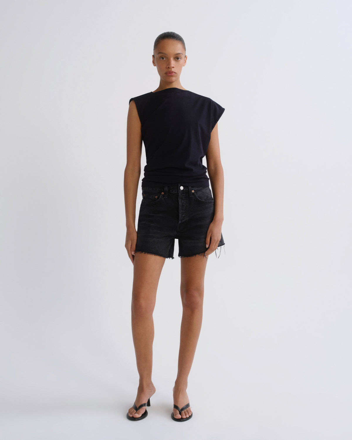 A model stands against a light gray studio background wearing a dark navy sleeveless top with asymmetrical shoulder cutouts, black denim shorts, and black thong sandals.