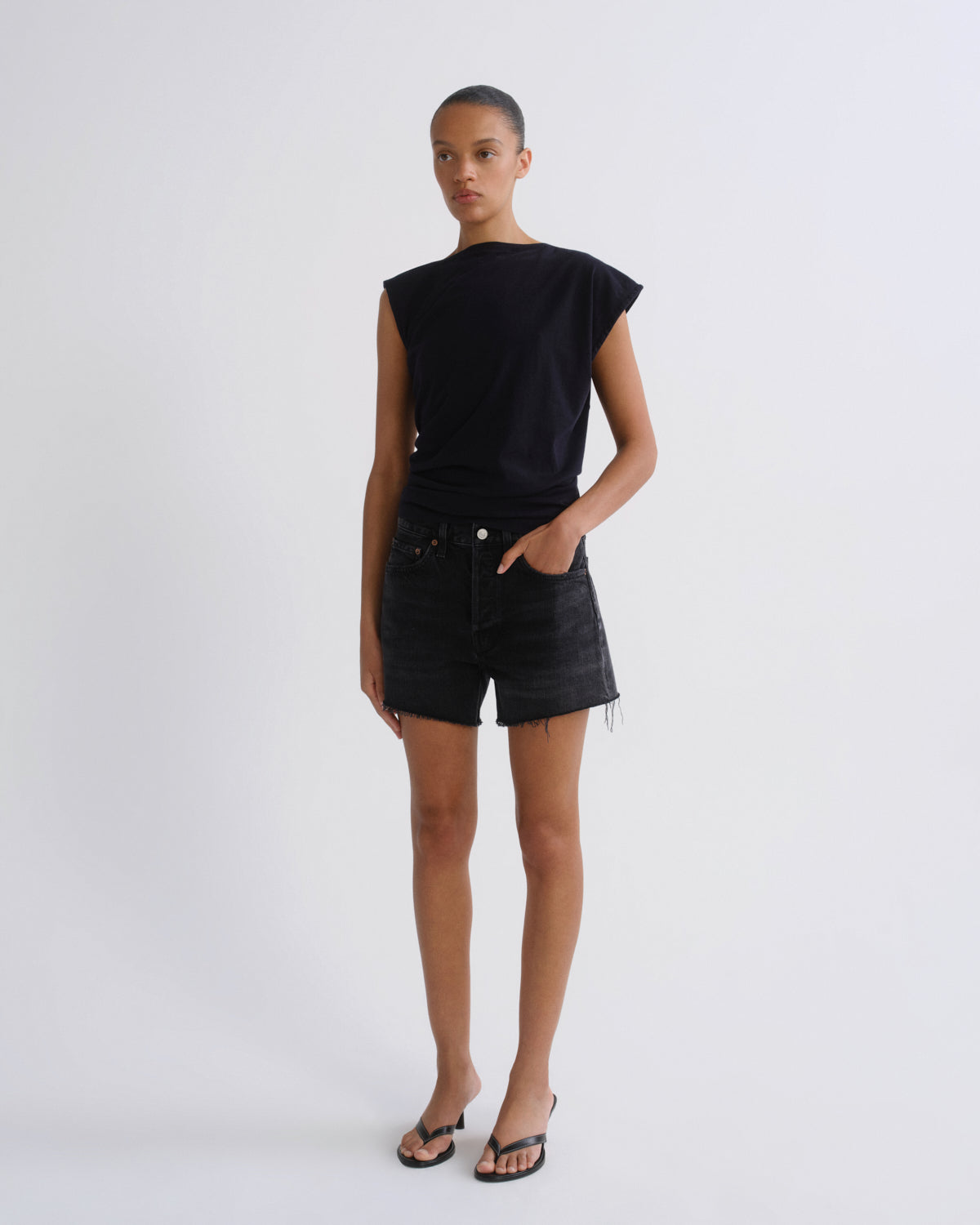 A model stands against a light gray studio background wearing a black sleeveless top with ruffled shoulders, black denim shorts, and black thong sandals.