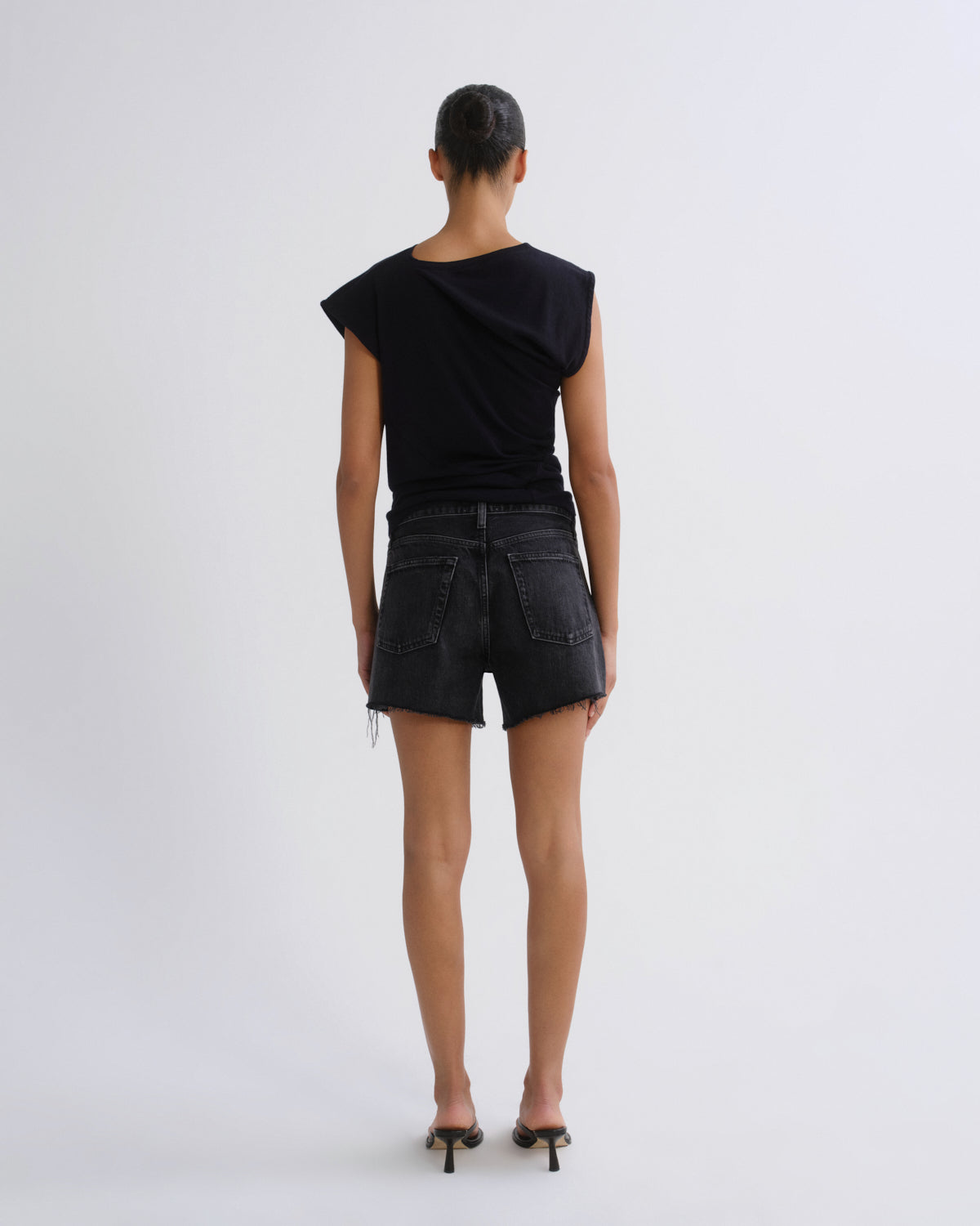 A model viewed from the back wearing a black asymmetrical short-sleeve top and black denim shorts, paired with black high-heeled sandals, against a plain light gray studio background.