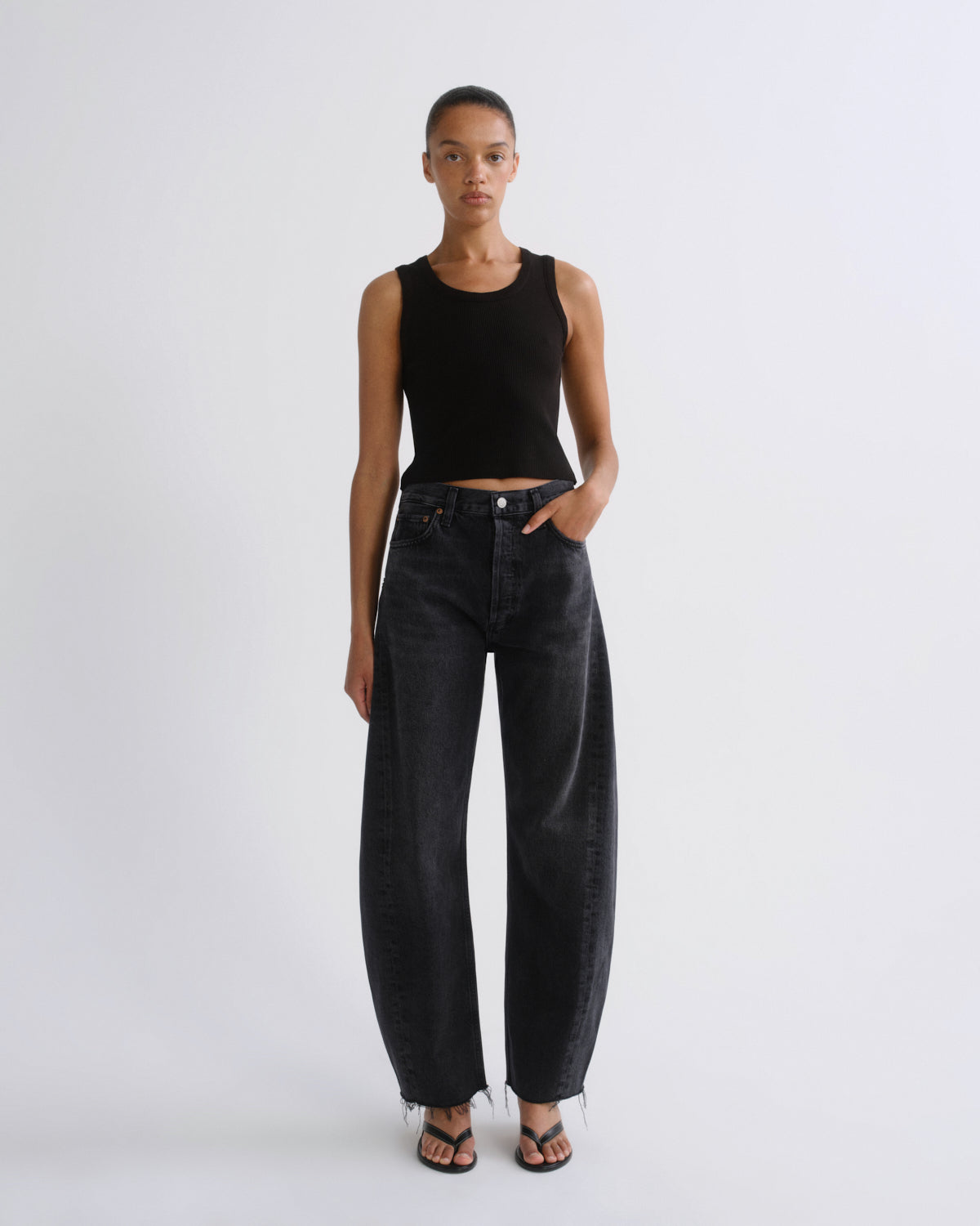 A model stands against a light gray studio background wearing a black ribbed sleeveless crop top and wide-leg dark gray jeans with fringe hems, paired with black sandals.