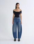 A model stands against a light gray studio background wearing a black off-the-shoulder top, wide-leg blue jeans, and black pointed shoes.