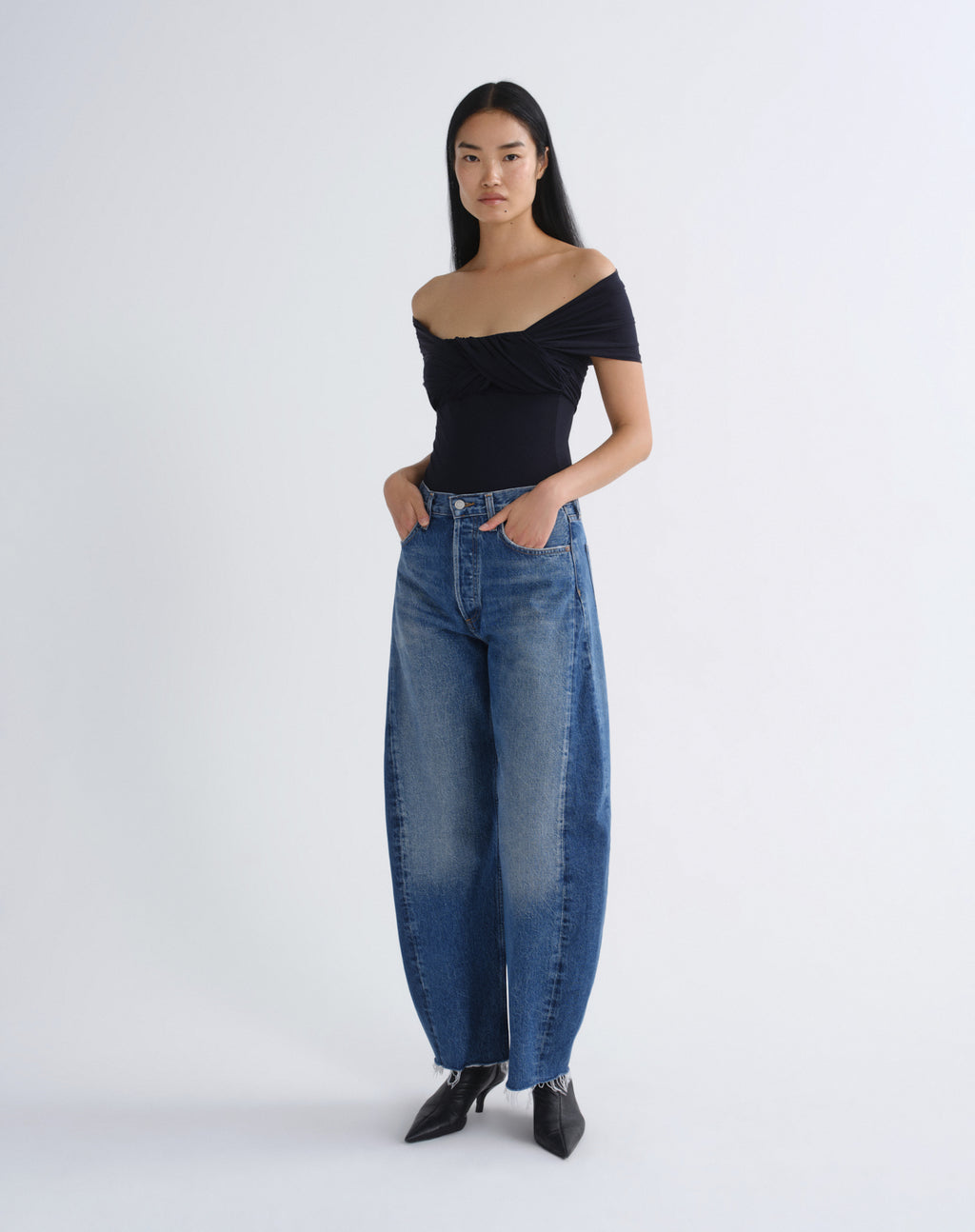 A model stands against a light gray studio background wearing a black off-the-shoulder top, wide-leg blue jeans, and black pointed shoes.