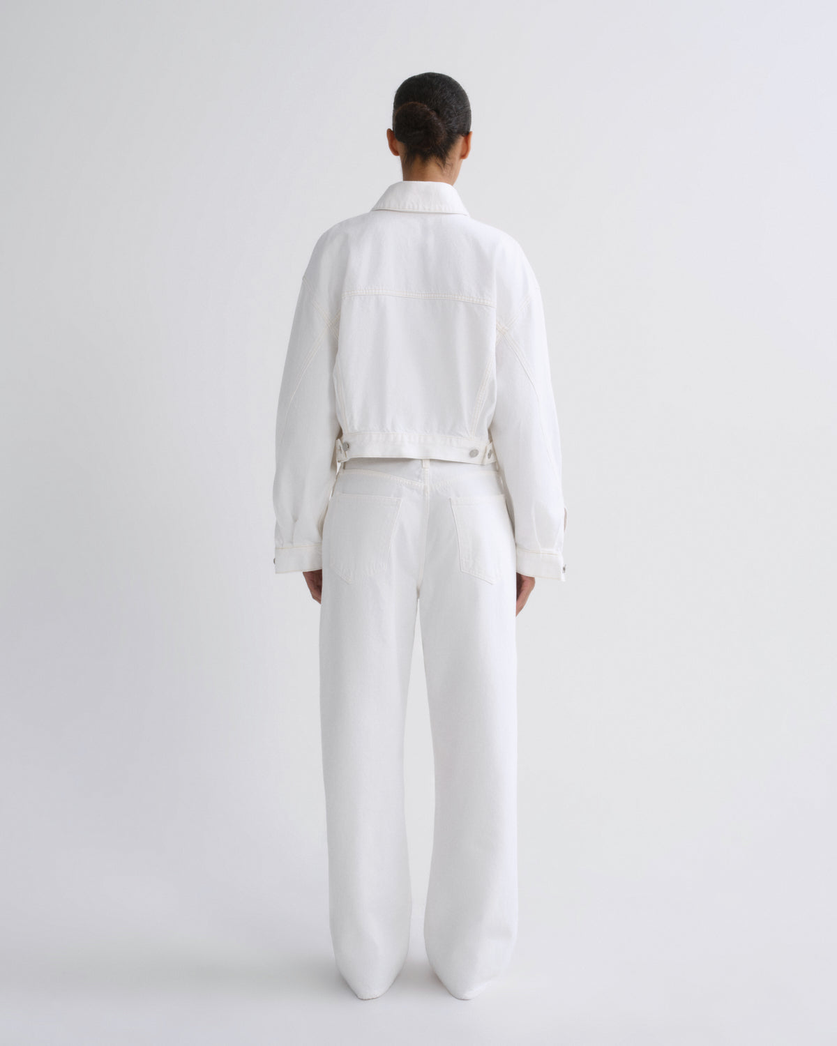 A model viewed from the back wearing a white cropped jacket and matching white wide-leg trousers against a plain light gray studio background.