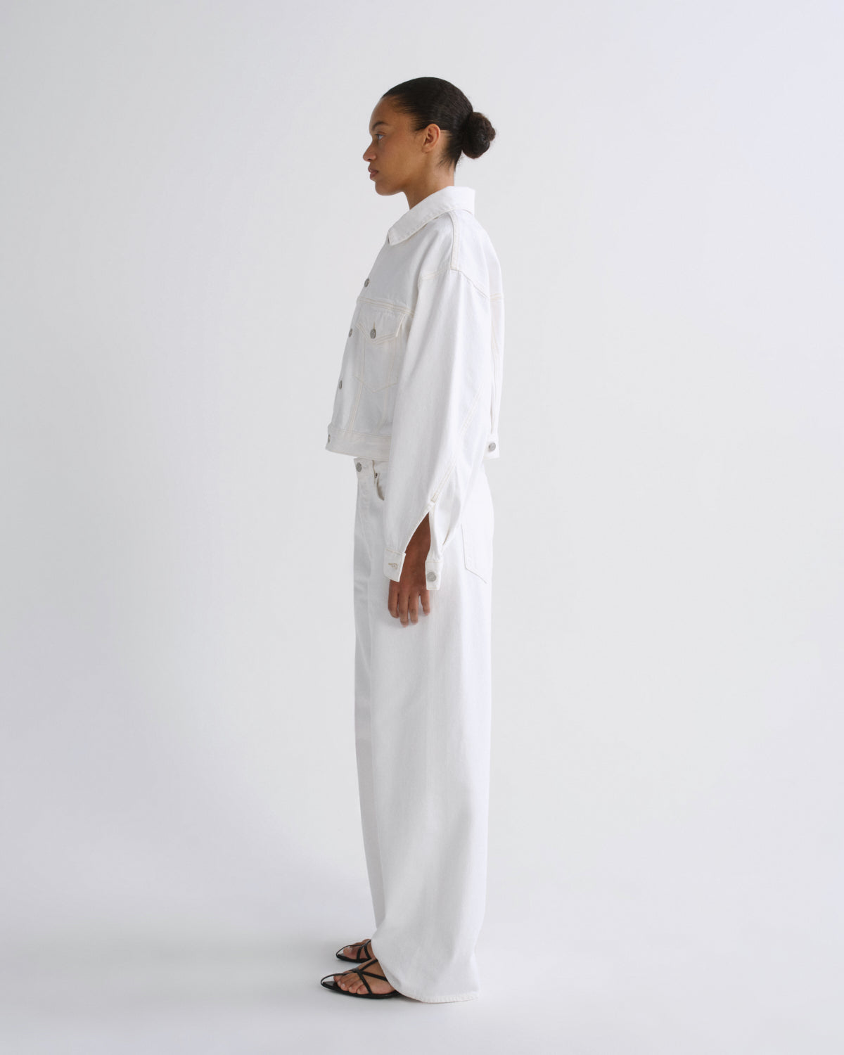 A model in profile wearing a white cropped denim jacket and matching wide-leg trousers, standing against a plain white studio background.