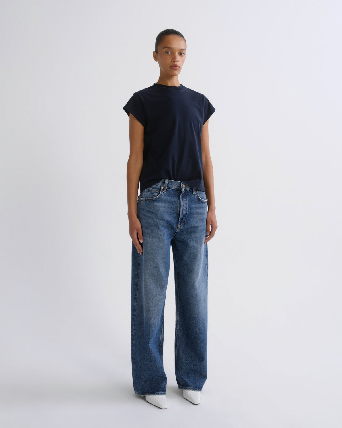 A model stands against a light gray studio background wearing a dark navy short-sleeve t-shirt, blue wide-leg jeans, and white pointed shoes.