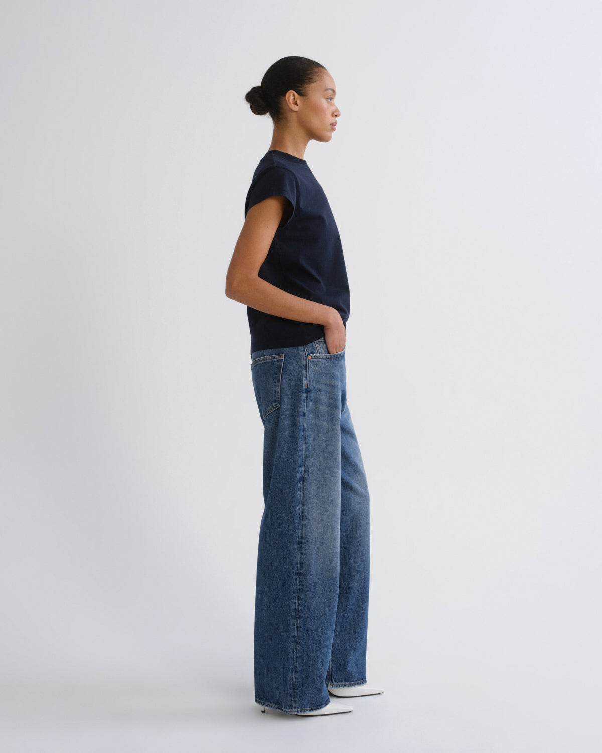 A model standing in profile wearing a dark navy short-sleeve top, wide-leg blue jeans, and white sneakers against a plain light gray studio background.