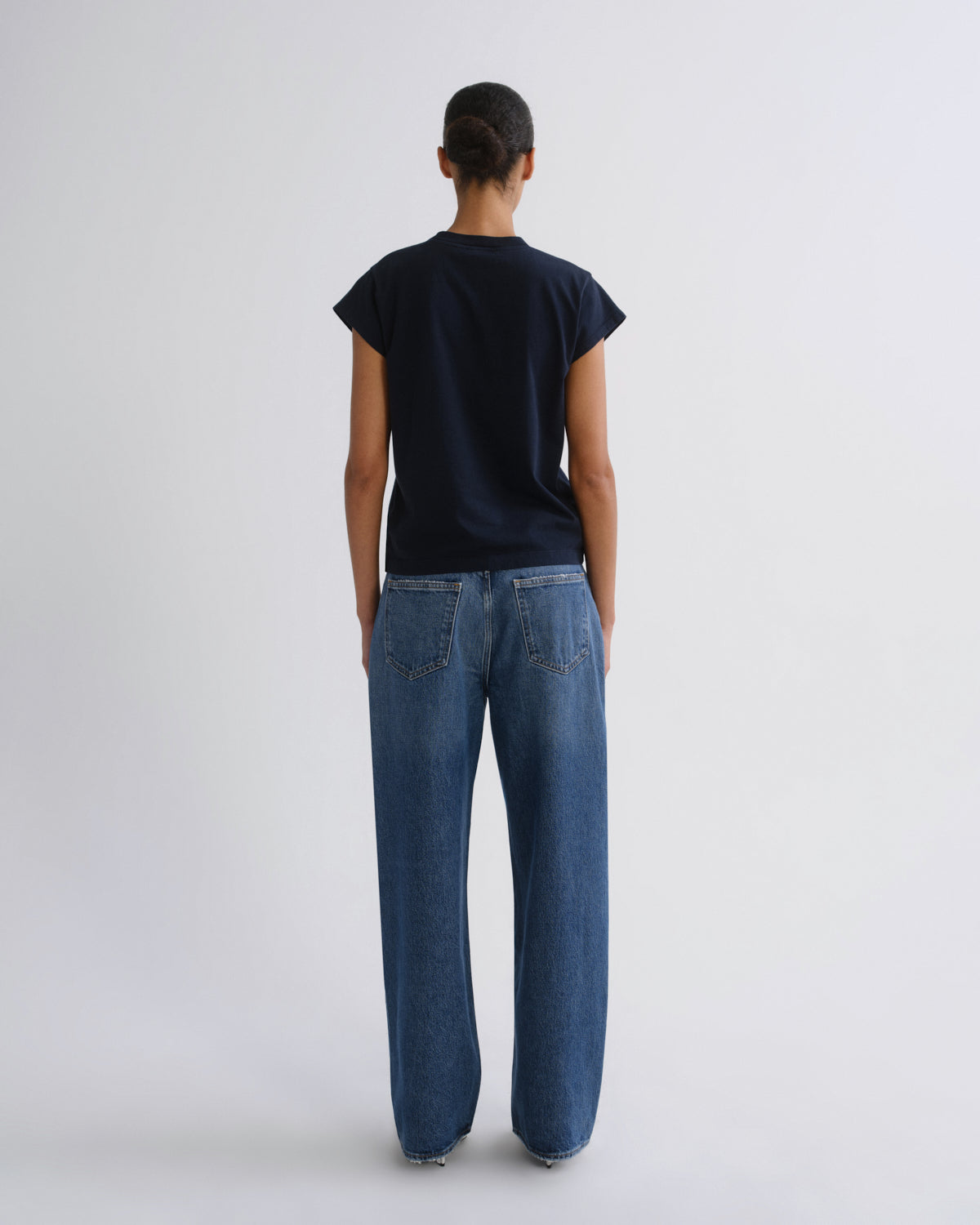 A model viewed from behind wearing a dark navy blue short-sleeve t-shirt and wide-leg blue denim jeans, standing against a light gray studio background.