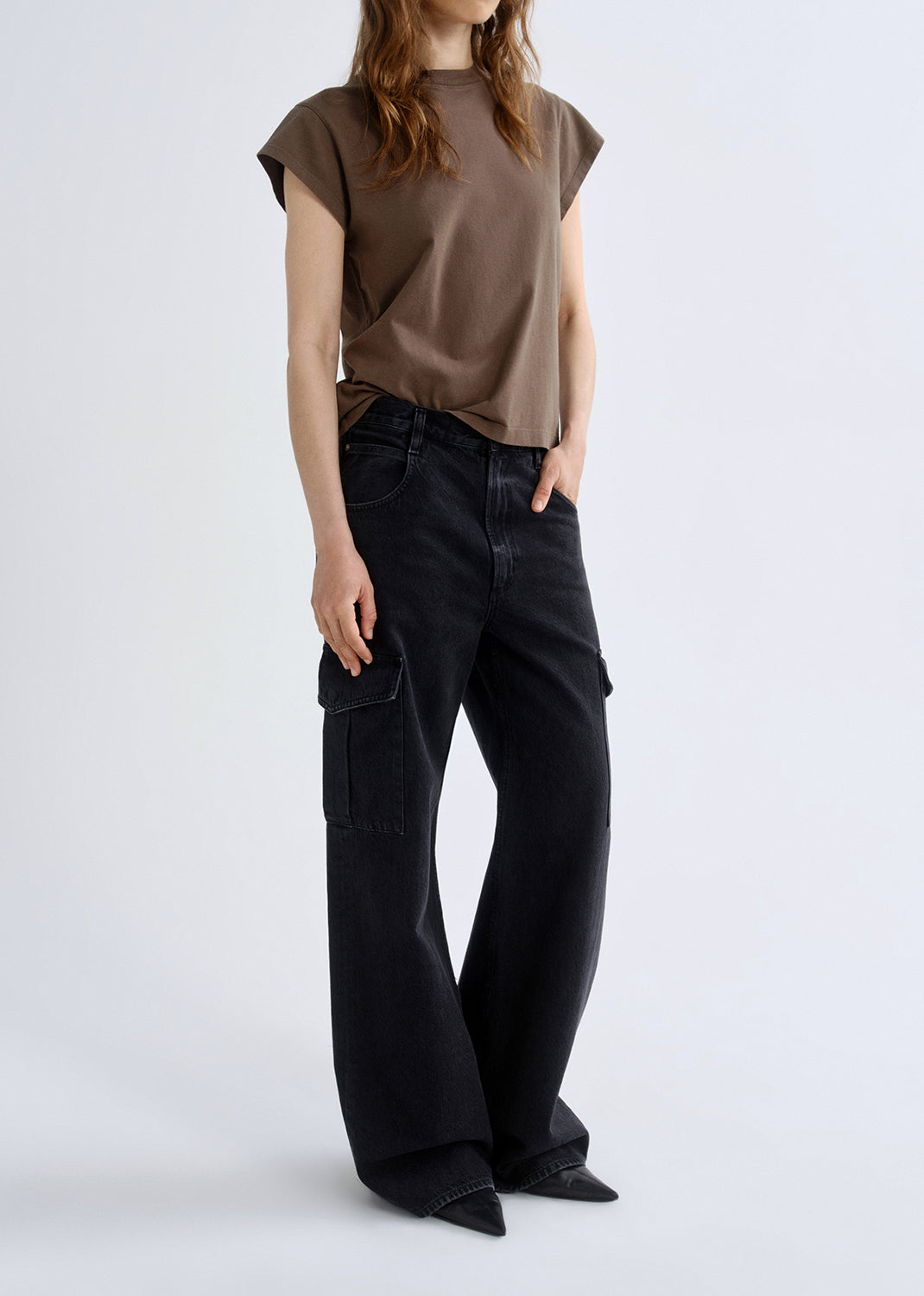 Person wearing a brown cap sleeve tee with black wide leg jeans viewed from the side on a grey background.