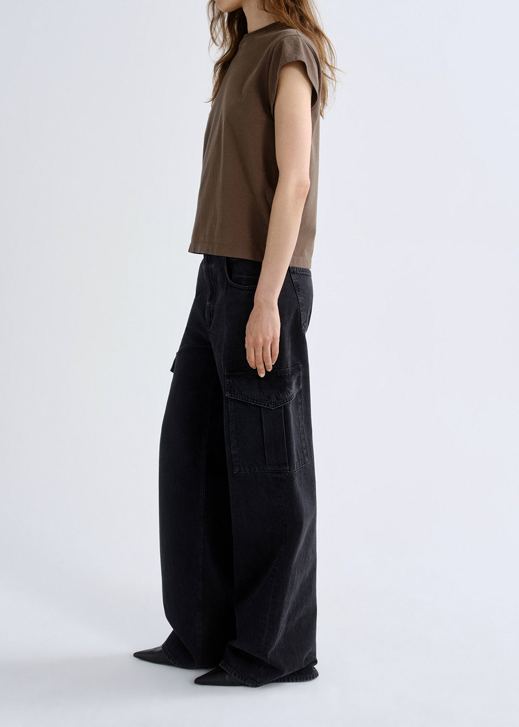 Person wearing a brown cap sleeve tee with black wide leg jeans viewed from the side on a grey background.