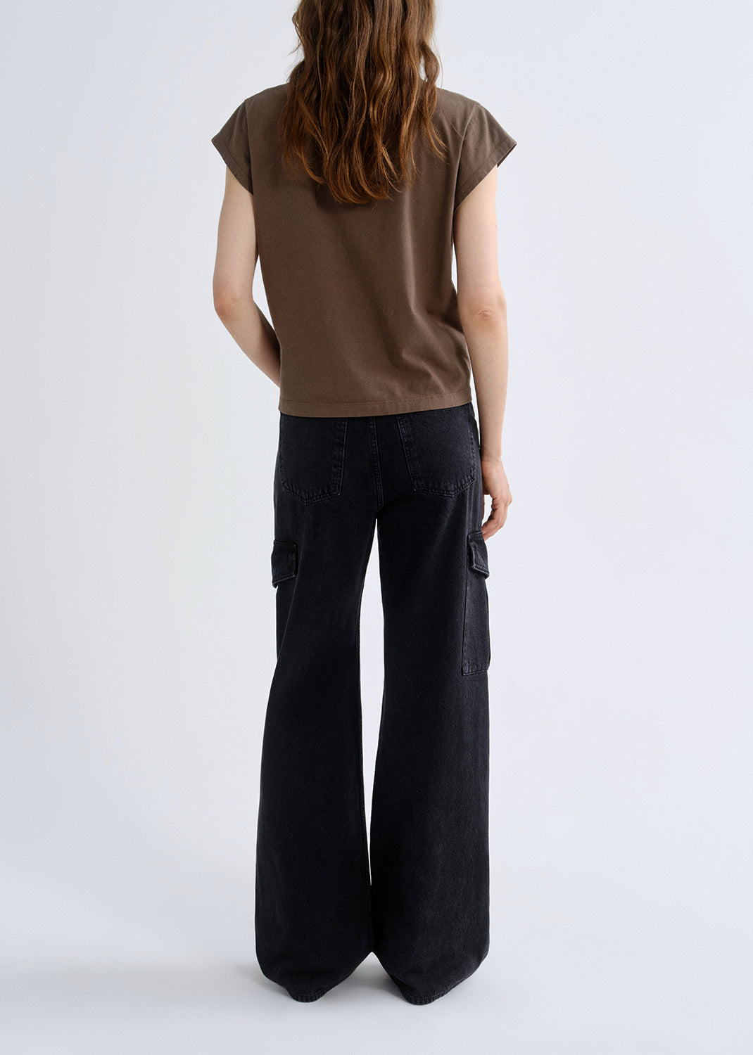Person wearing a brown cap sleeve tee with black wide leg jeans viewed from the back on a grey background.