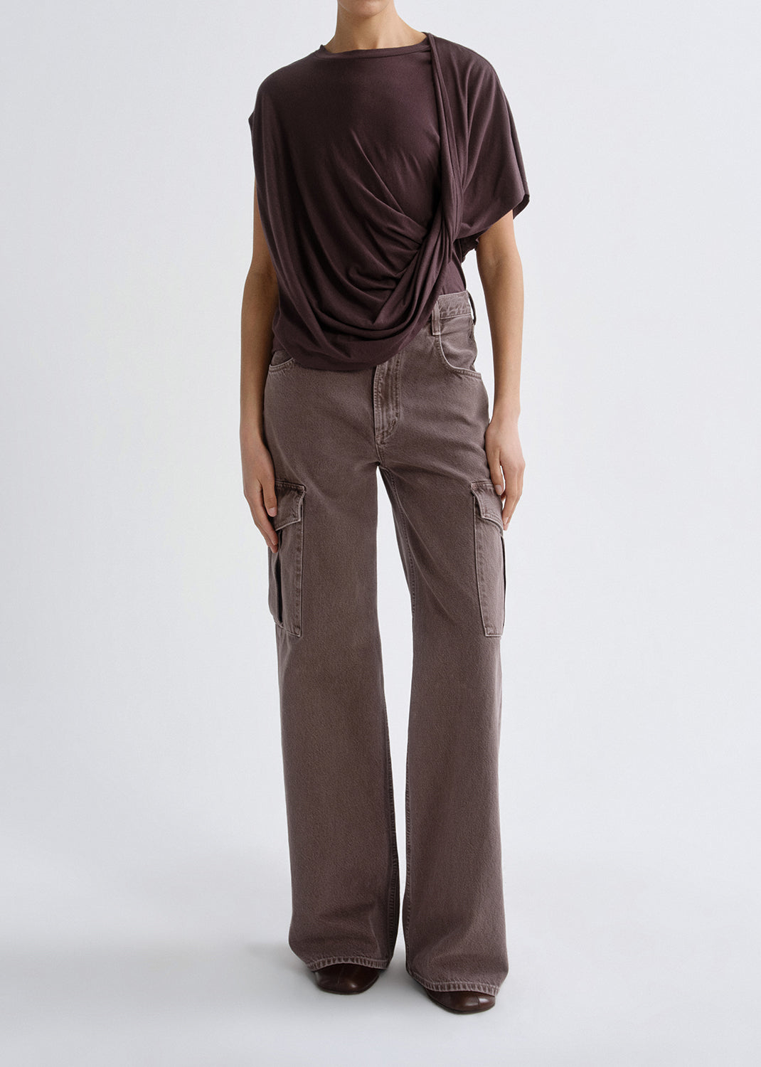 A model wearing a dark brown draped short-sleeve top and wide-leg cargo pants in a muted taupe-brown color, photographed against a grey background.